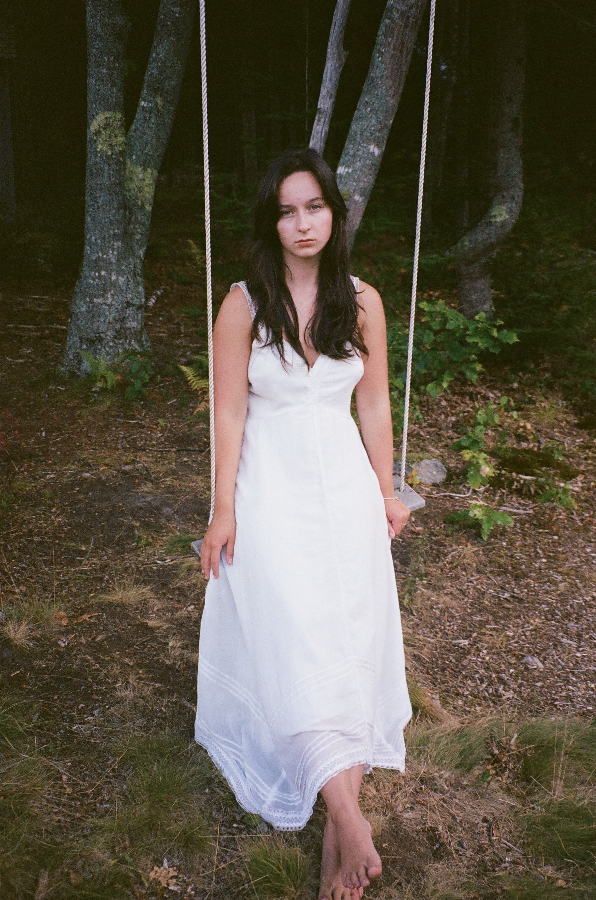 A young woman in a white dress sitting on a swing in a wooded area.