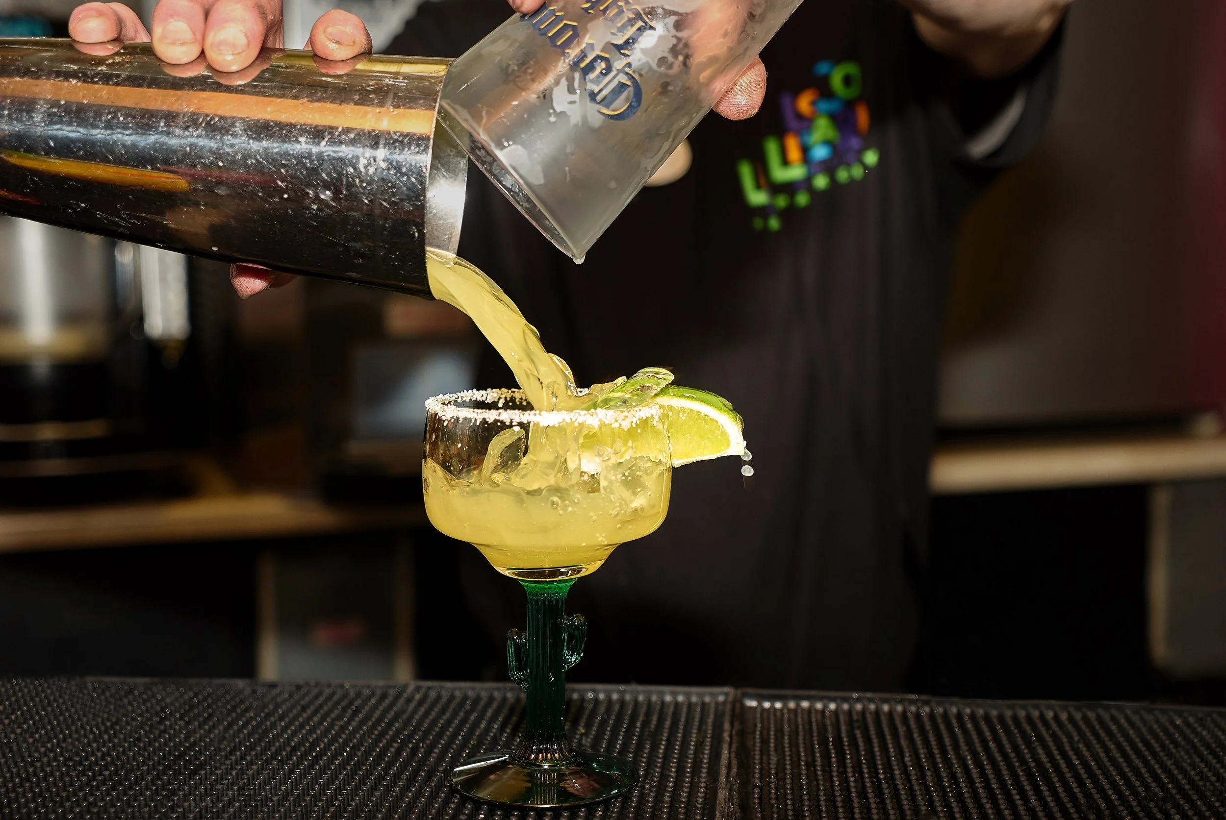 A bartender pours yellow cocktail from a shaker into a glass garnished with lime, with salt rim.