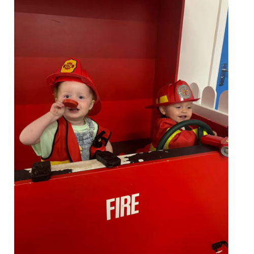 Two young children dressed as firefighters sitting in a toy fire truck. The girl on the left has a red firefighter hat and vest, smiling and holding a plastic tool. The boy on the right, wearing a firefighter helmet and red jacket, is sitting behind the steering wheel.