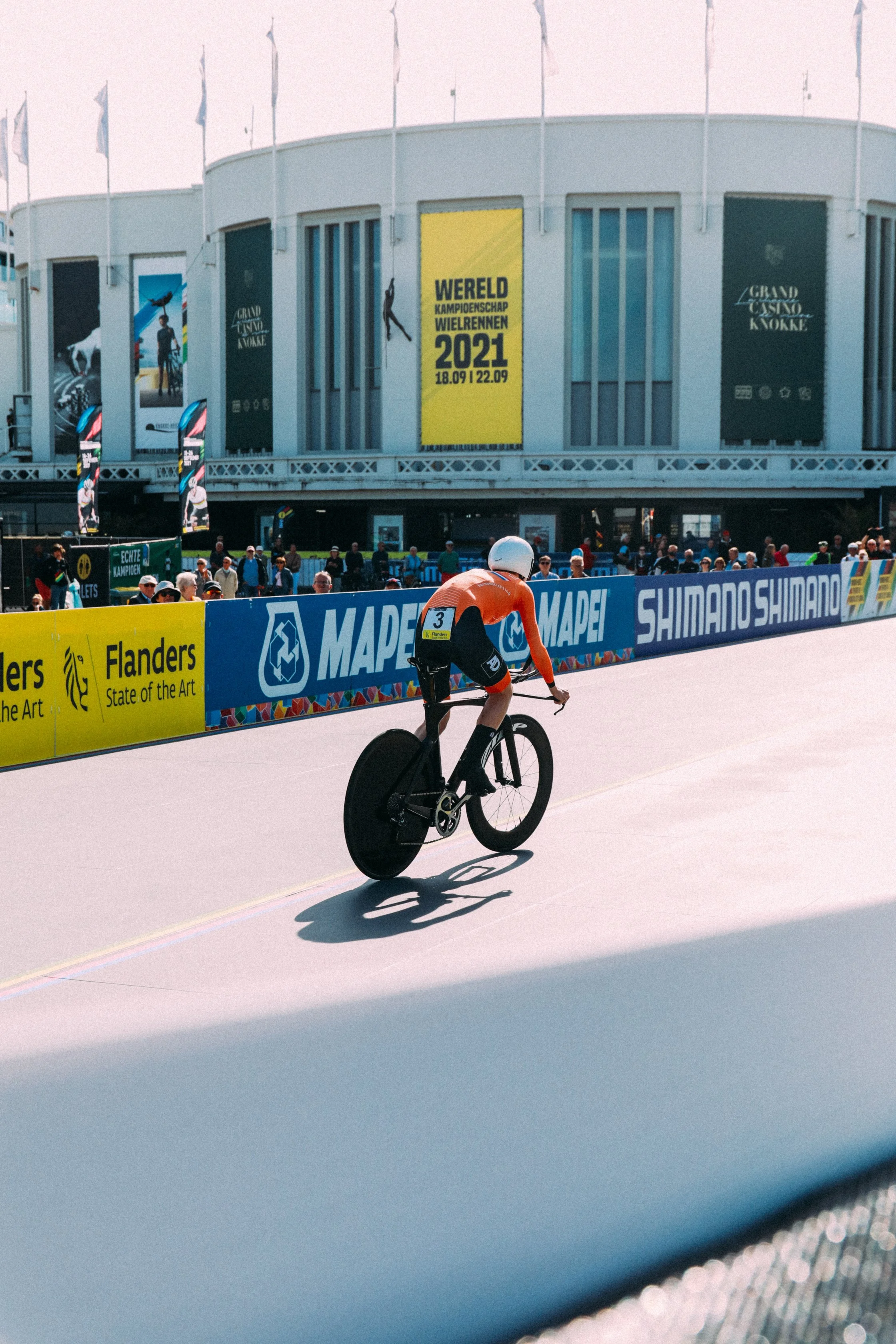 Cyclist in orange jersey racing at 2021 World Cycling Championships, venue with banners, and spectators in background.