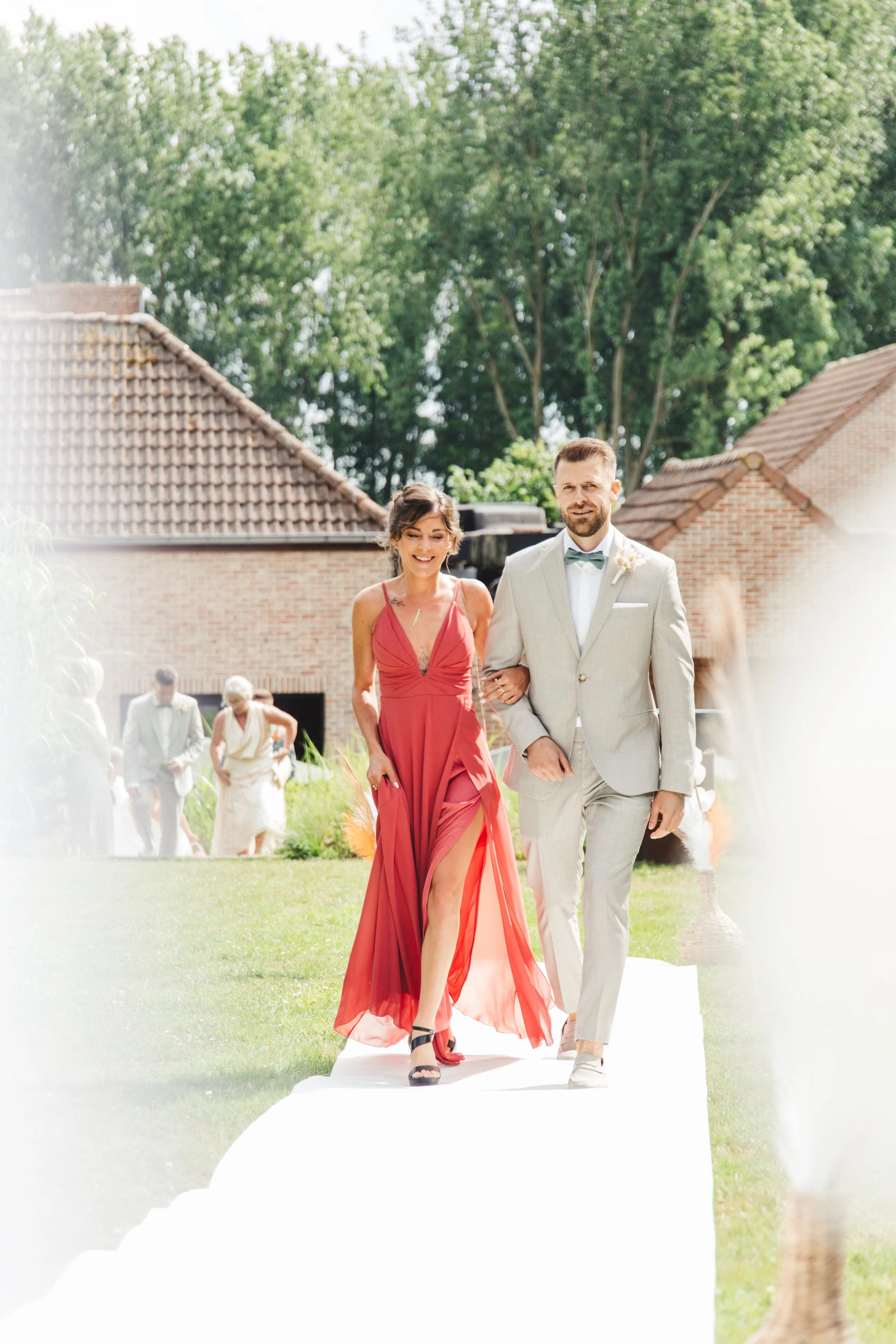 A woman in a red dress and a man in a light gray suit walking down an outdoor aisle, possibly during a wedding ceremony.