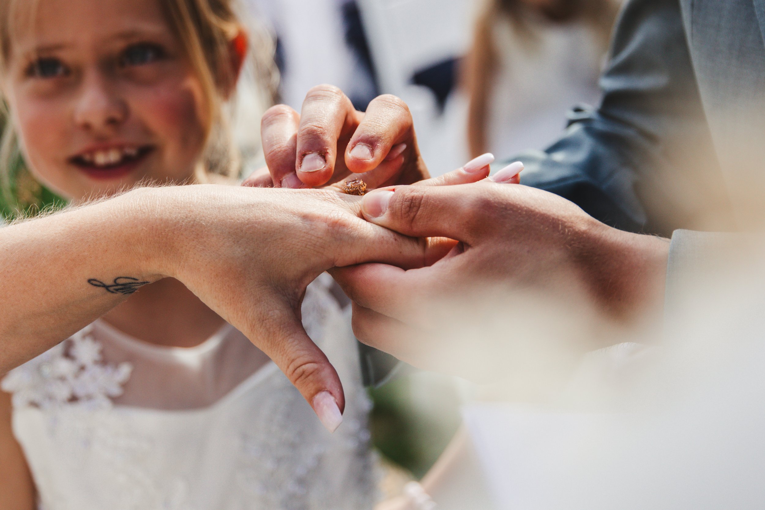 Close-up of hands exchanging rings at a wedding ceremony, with a young girl smiling in the background.