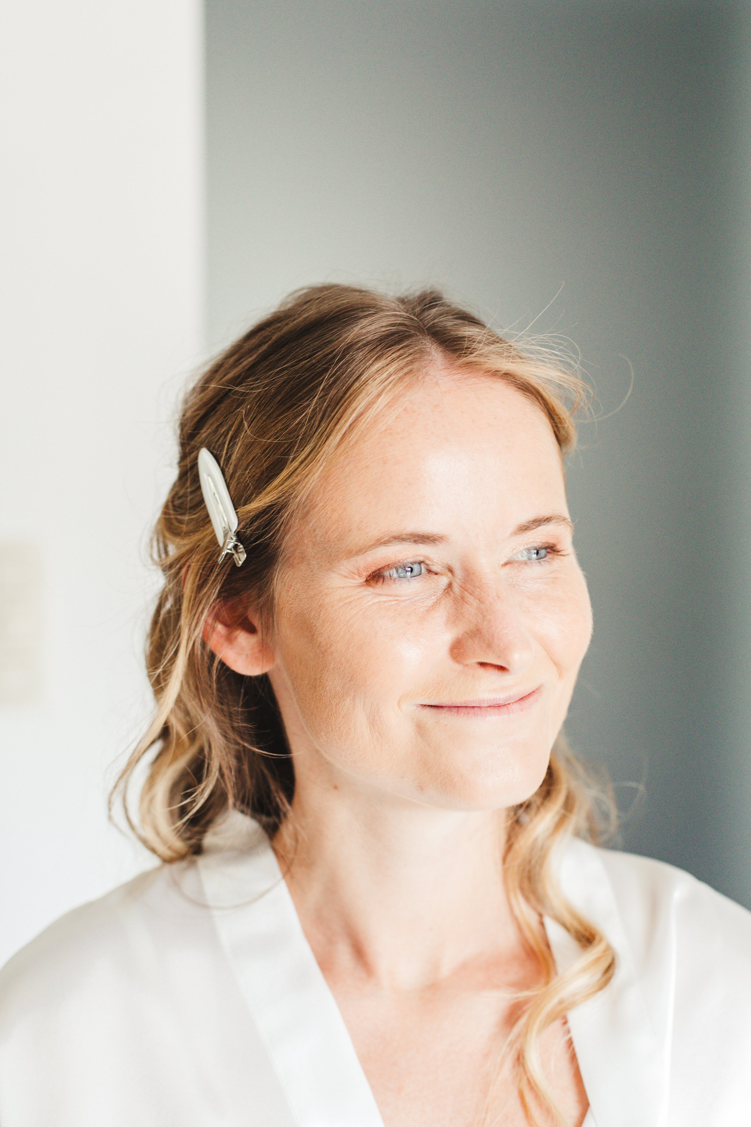 A woman with blonde hair wearing a white robe, smiling and looking sideways. Her hair is styled with a barrette.