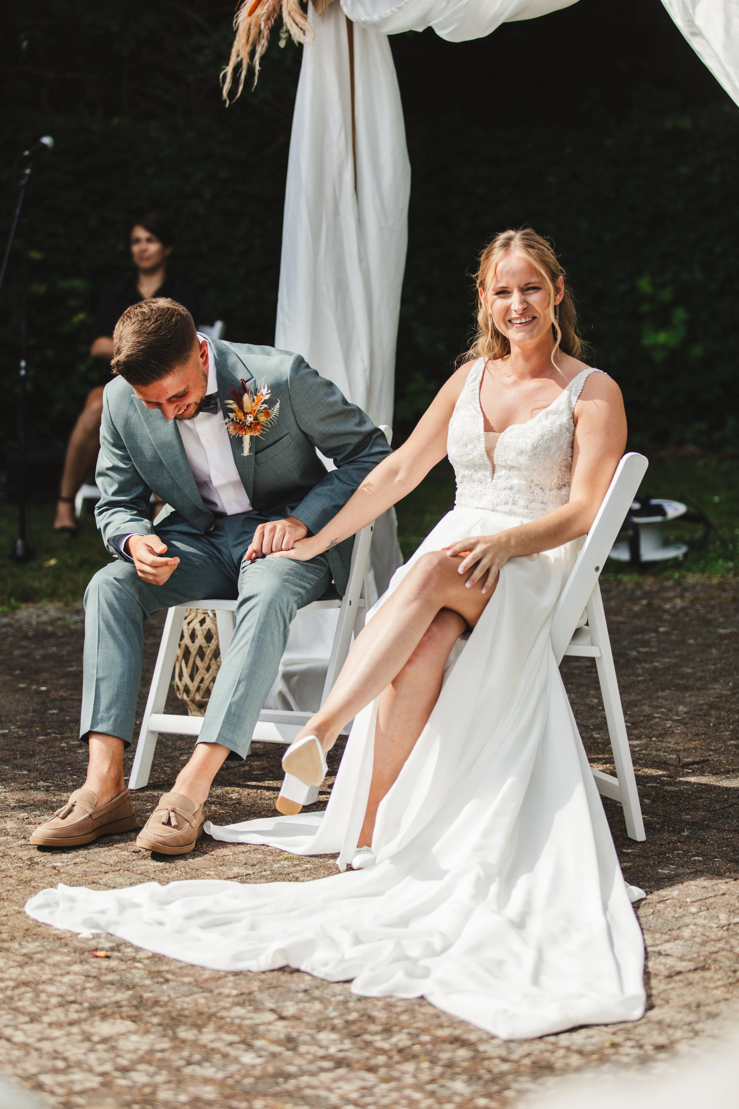 Bride and groom sitting outdoors holding hands, with the bride in a white dress and the groom in a light gray suit.