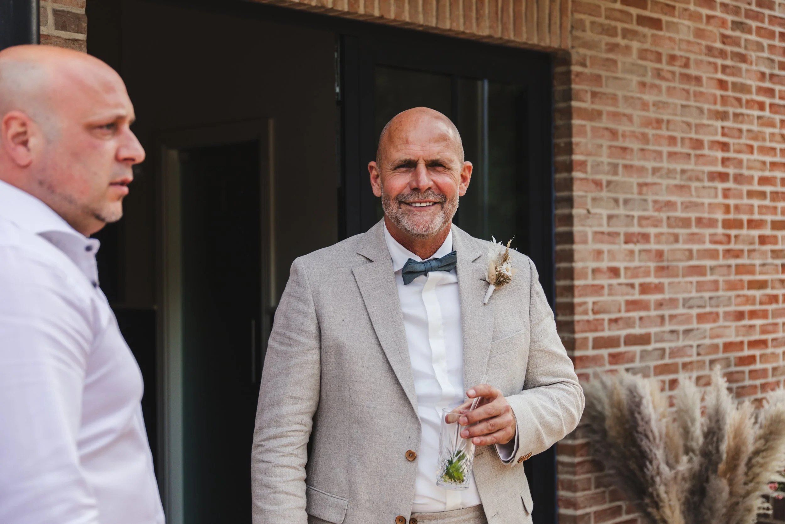 Two men at a formal event, one wearing a beige suit with a boutonniere, holding a drink, standing outside a brick building.