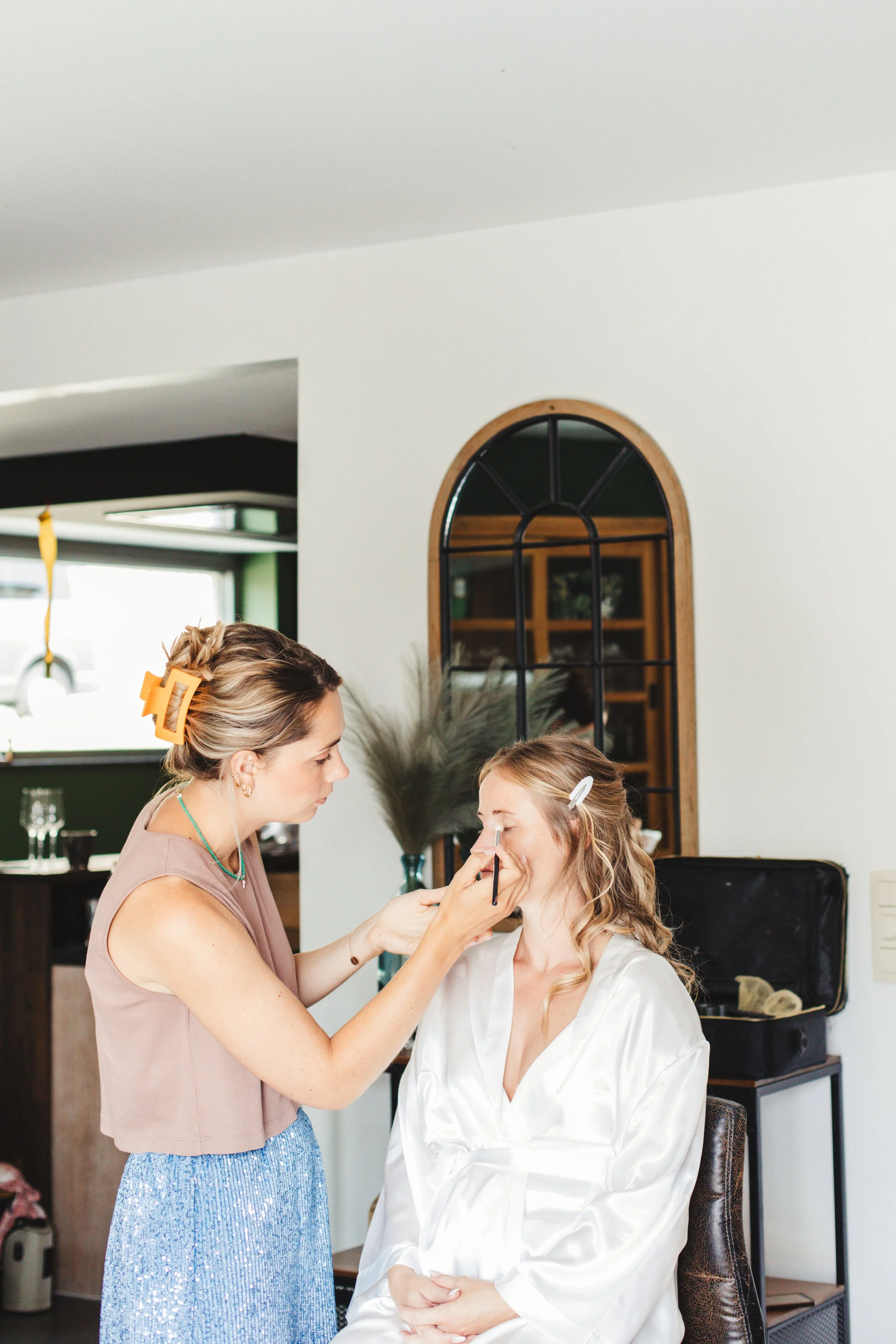 Woman getting makeup done by a makeup artist indoors, with decorative mirror and plant in background.