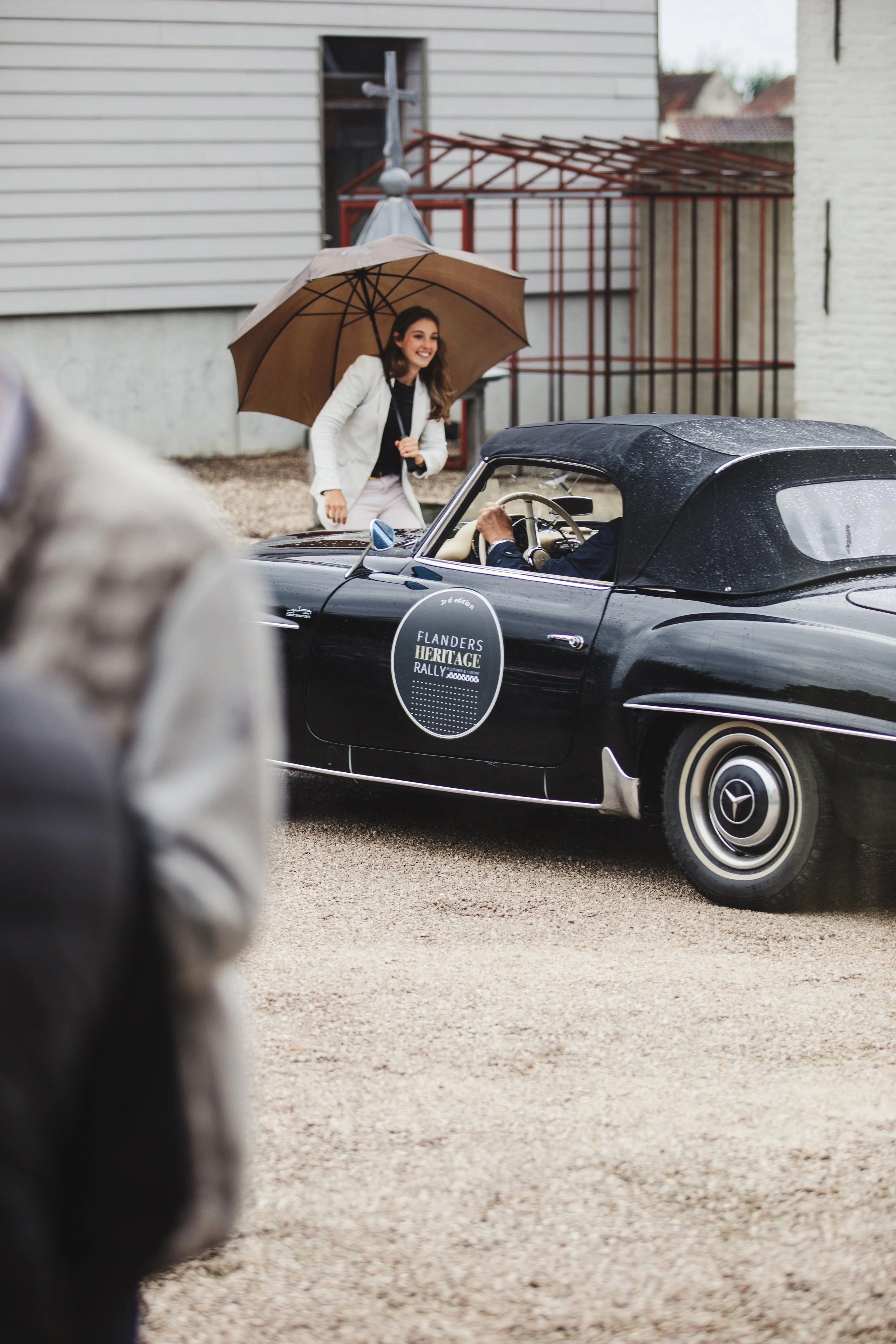 A smiling woman holding an umbrella stands next to a classic black car with "Flanders Heritage Rally" written on the door.