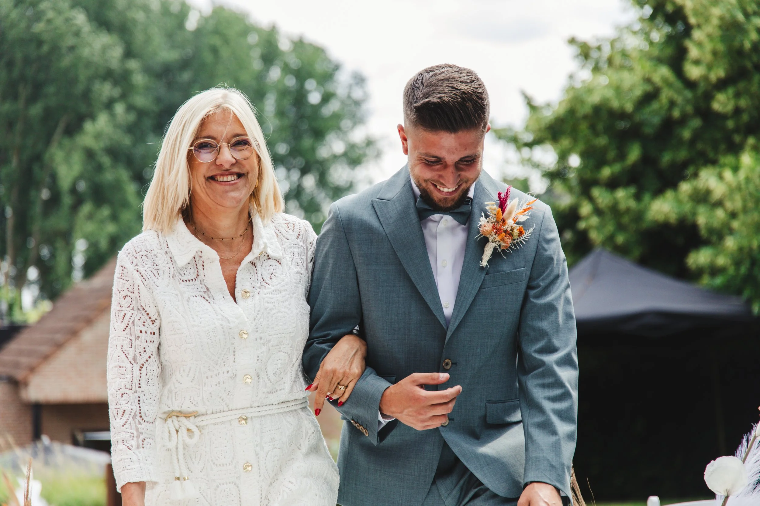 A smiling woman in a white lace dress and glasses walks arm-in-arm with a man in a light blue suit and bow tie, wearing a floral boutonniere, outdoors.