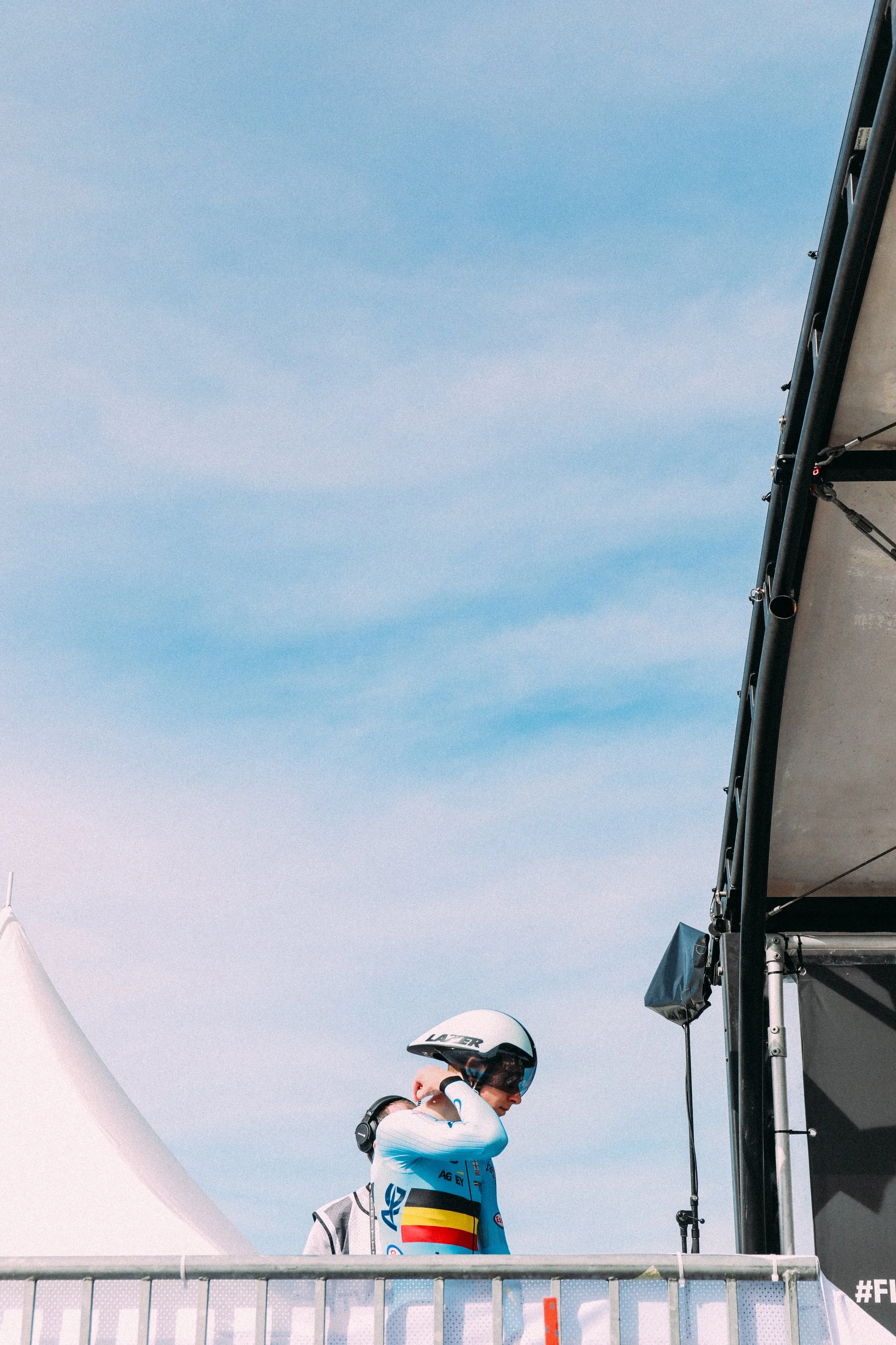 Cyclist wearing a helmet and racing suit preparing for a race near a tent and stage under a blue sky.