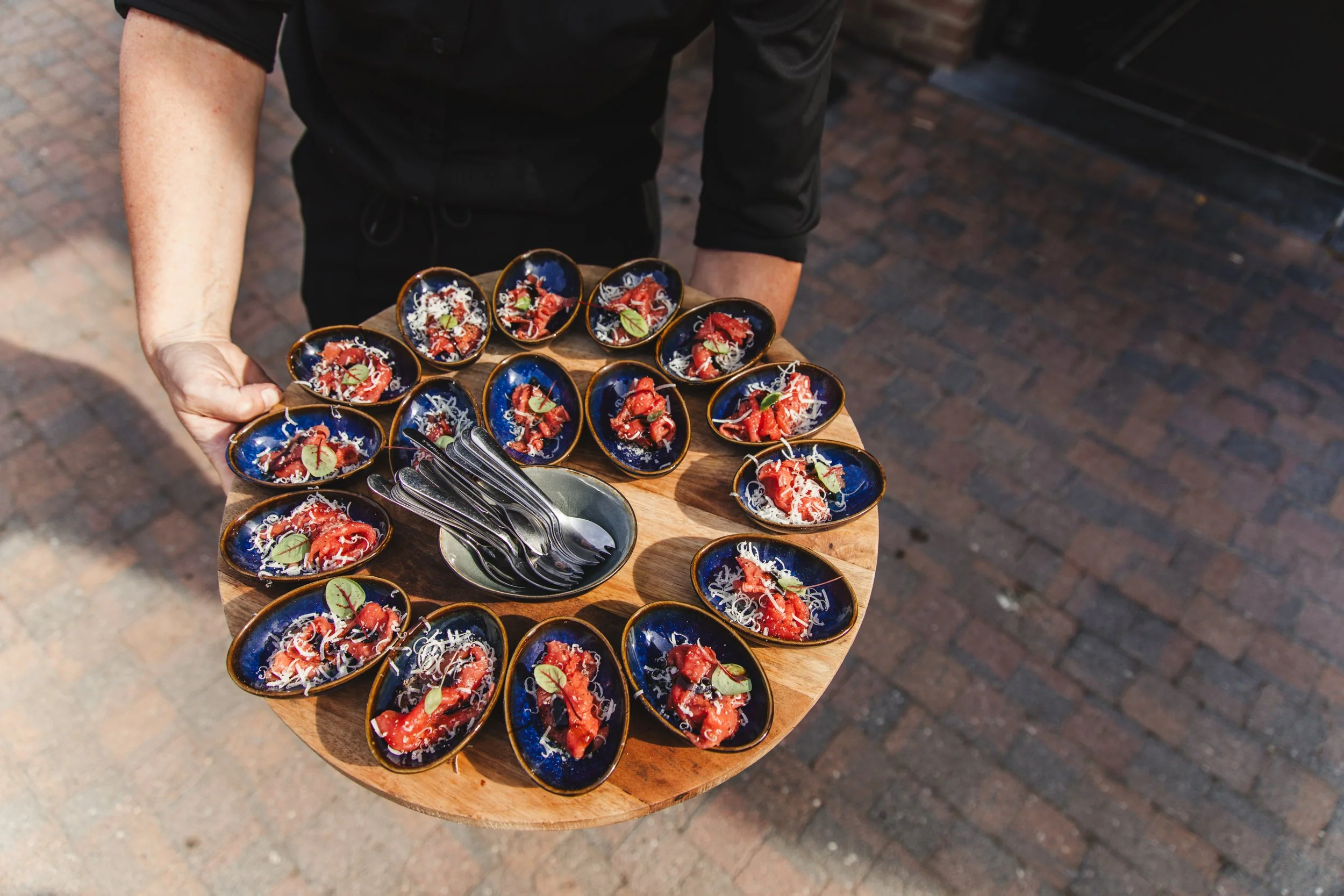 Person holding wooden tray with small dishes of food and a bowl of spoons.