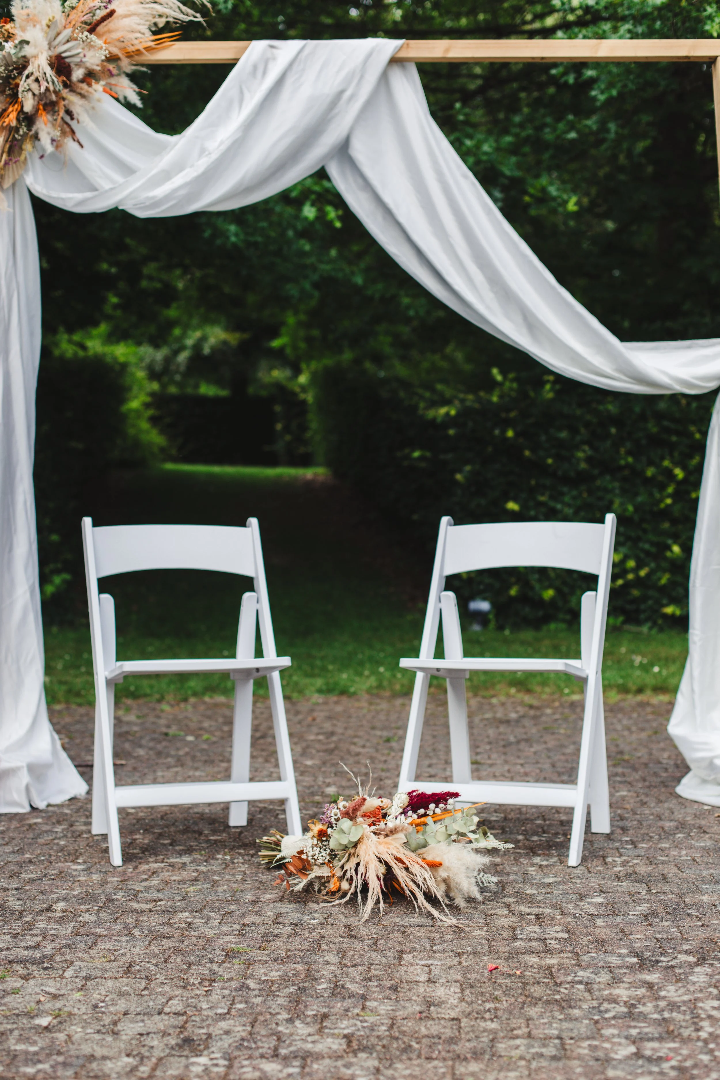 Outdoor wedding ceremony setup with two white chairs under an arch adorned with white fabric and dried flowers, on a brick pathway in a garden setting.