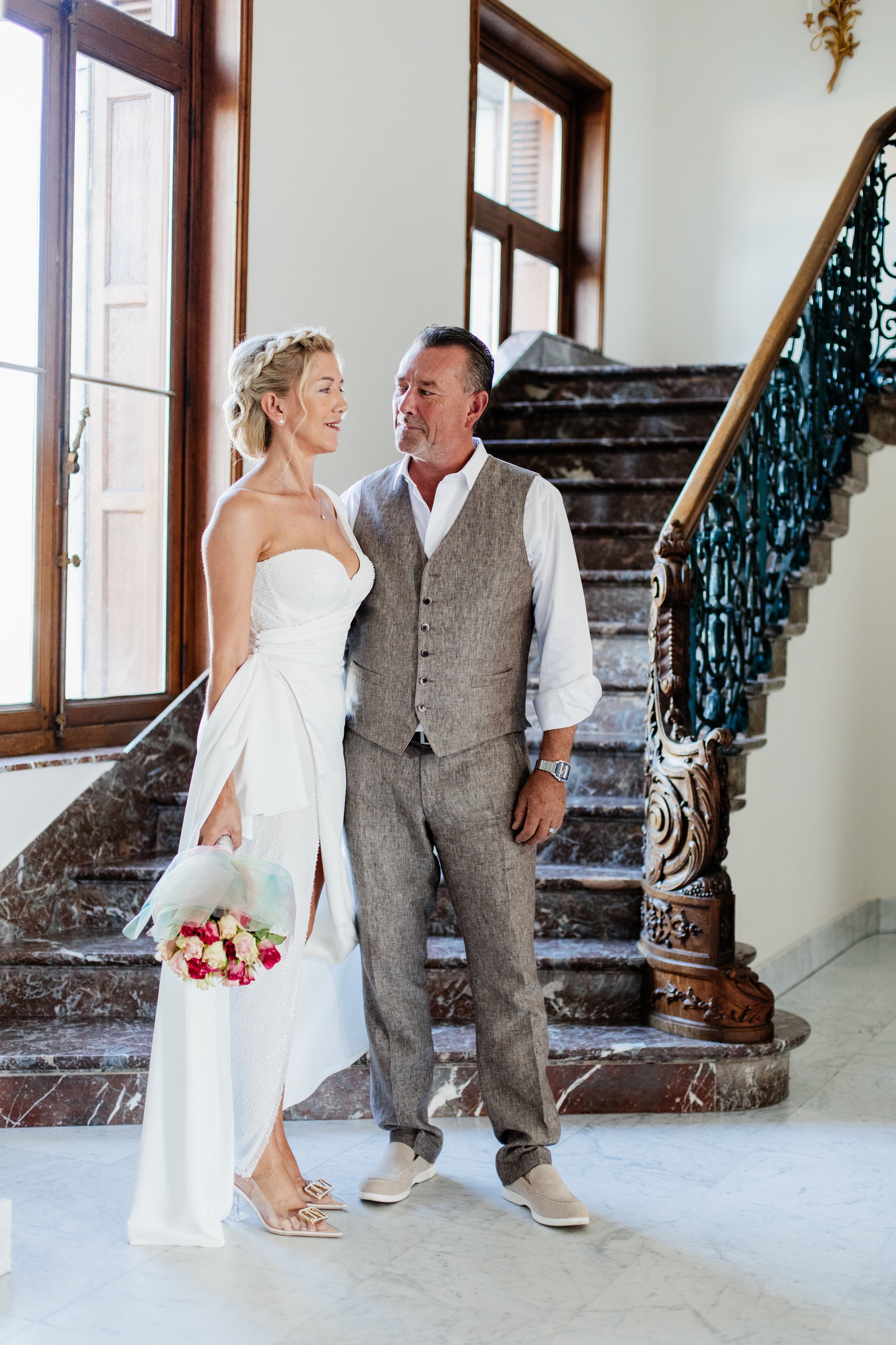 A couple stands facing each other in a formal setting. The woman, wearing a white dress and holding a bouquet of flowers, is smiling. The man is in a gray vest and pants. They are in front of a marble staircase with intricate railing.
