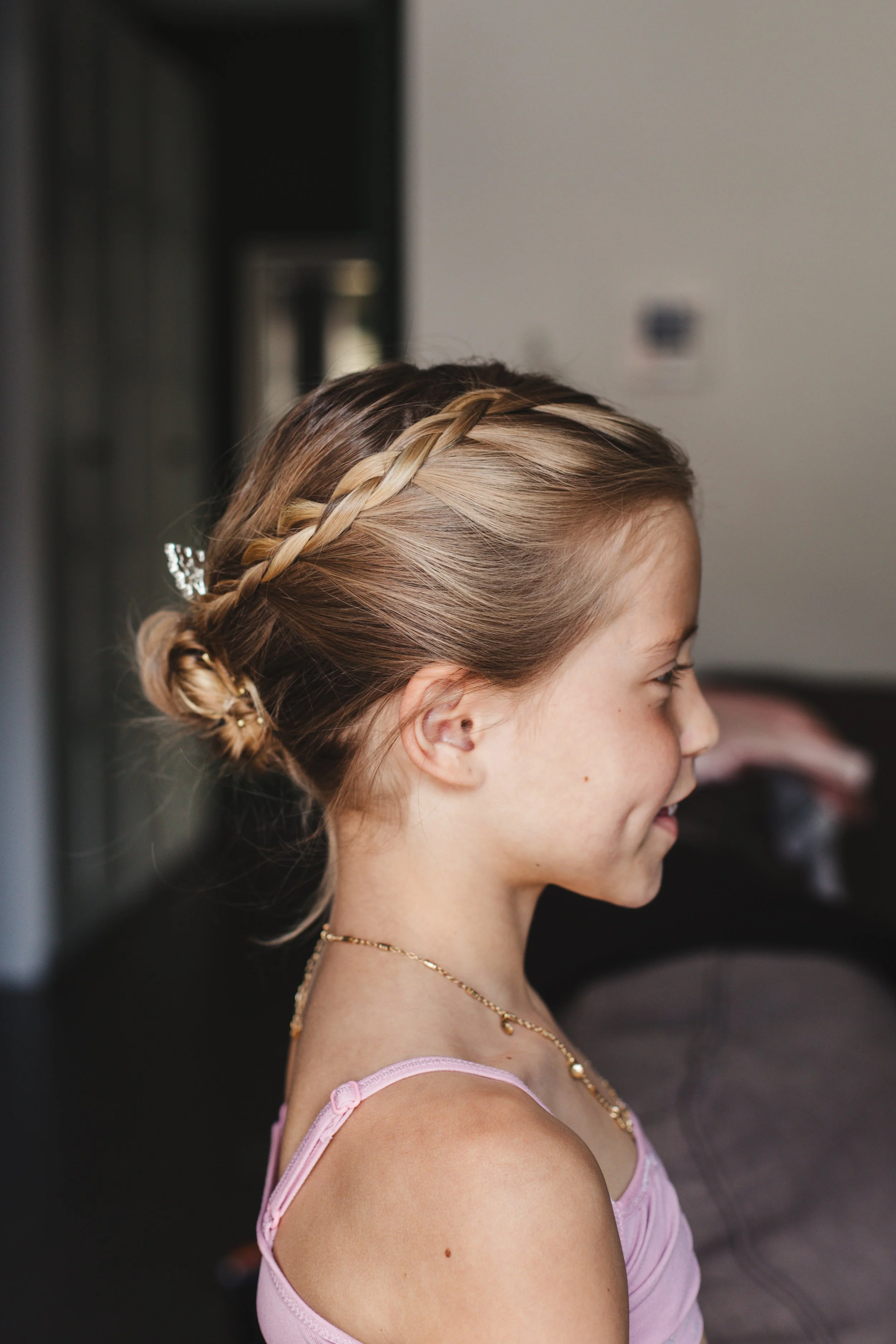A young girl with braided hair in a bun, wearing a pink top and gold necklace, profile view.