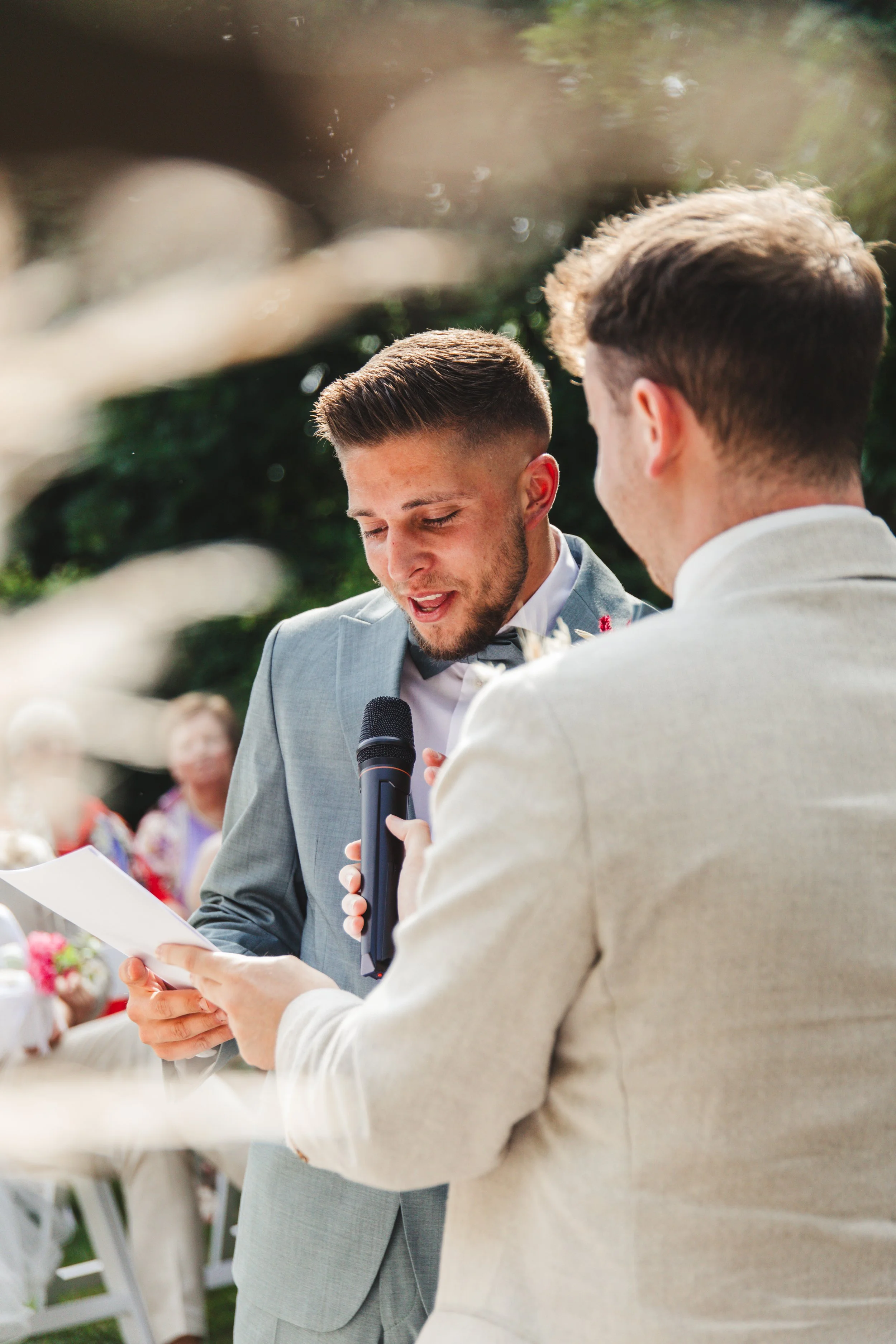 Two men in suits at an outdoor event, one holding a microphone and the other reading from a paper.