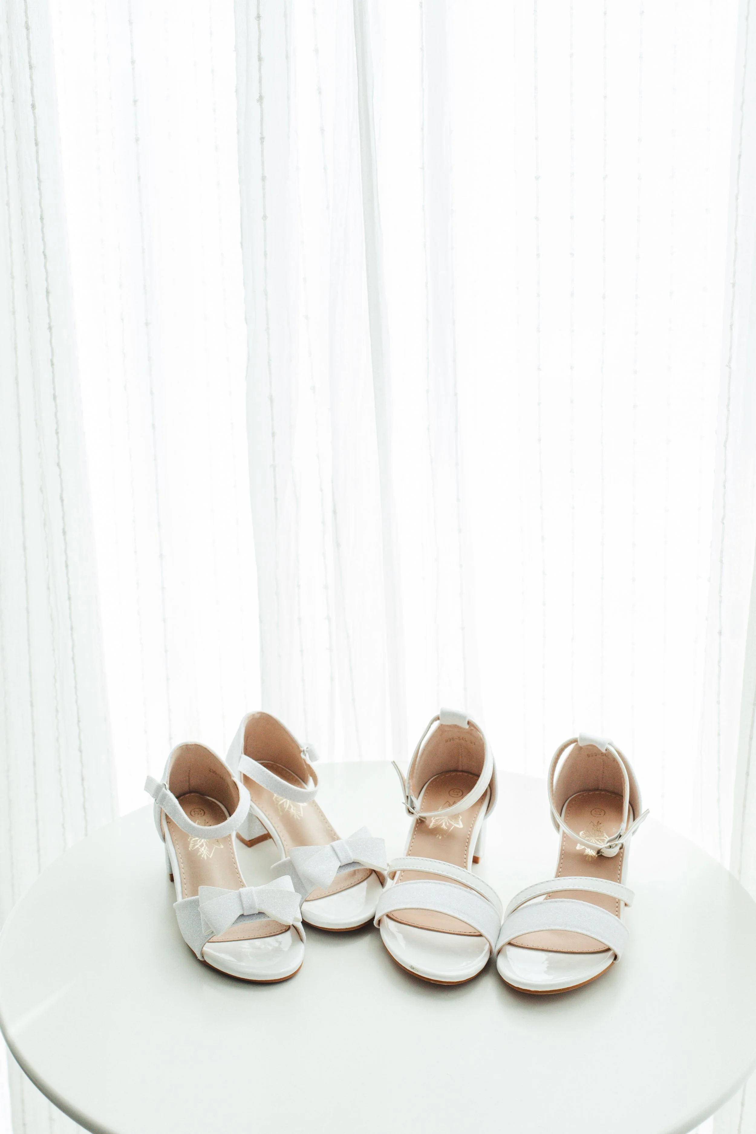 Two pairs of white high-heeled sandals with bows on a white table, against a sheer curtain background.