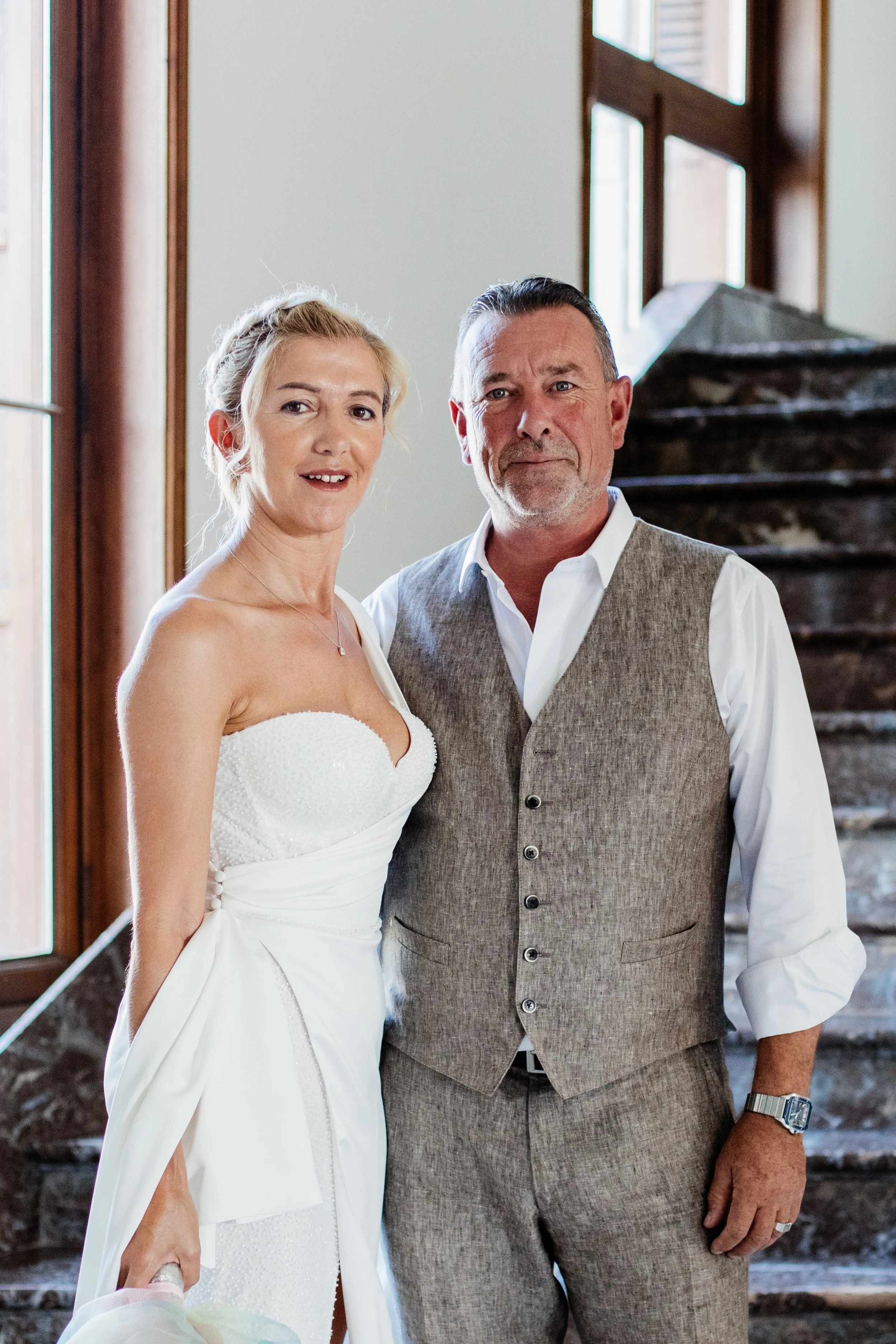A bride in a white dress stands next to a man in a beige vest and white shirt, both posing in front of a staircase indoors, with natural light coming through a window.