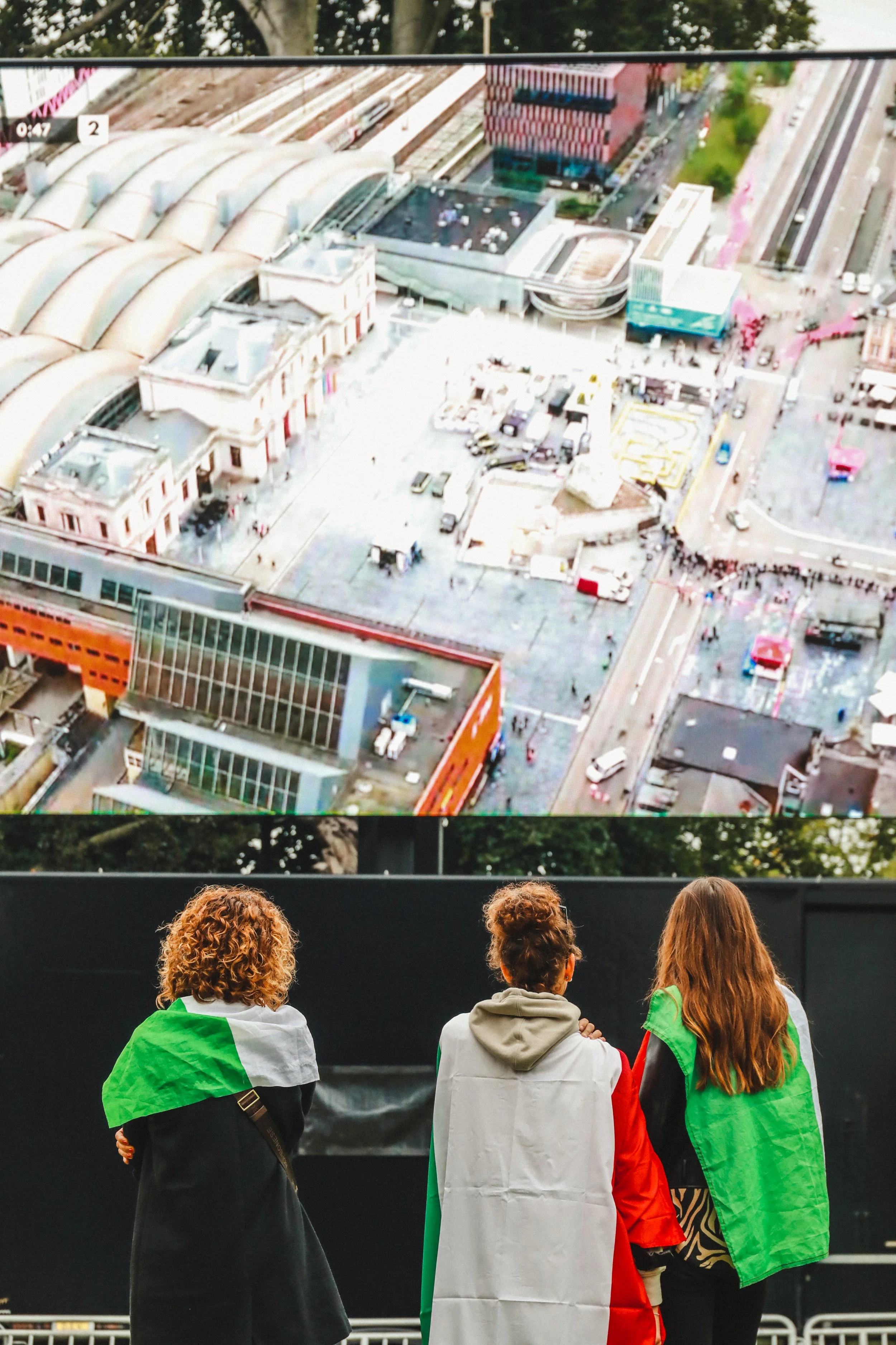 Three people wearing green, white, and red flags watch a large outdoor screen displaying an aerial view of a train station with nearby buildings and roads. The setting appears to be a public area with trees visible in the background.