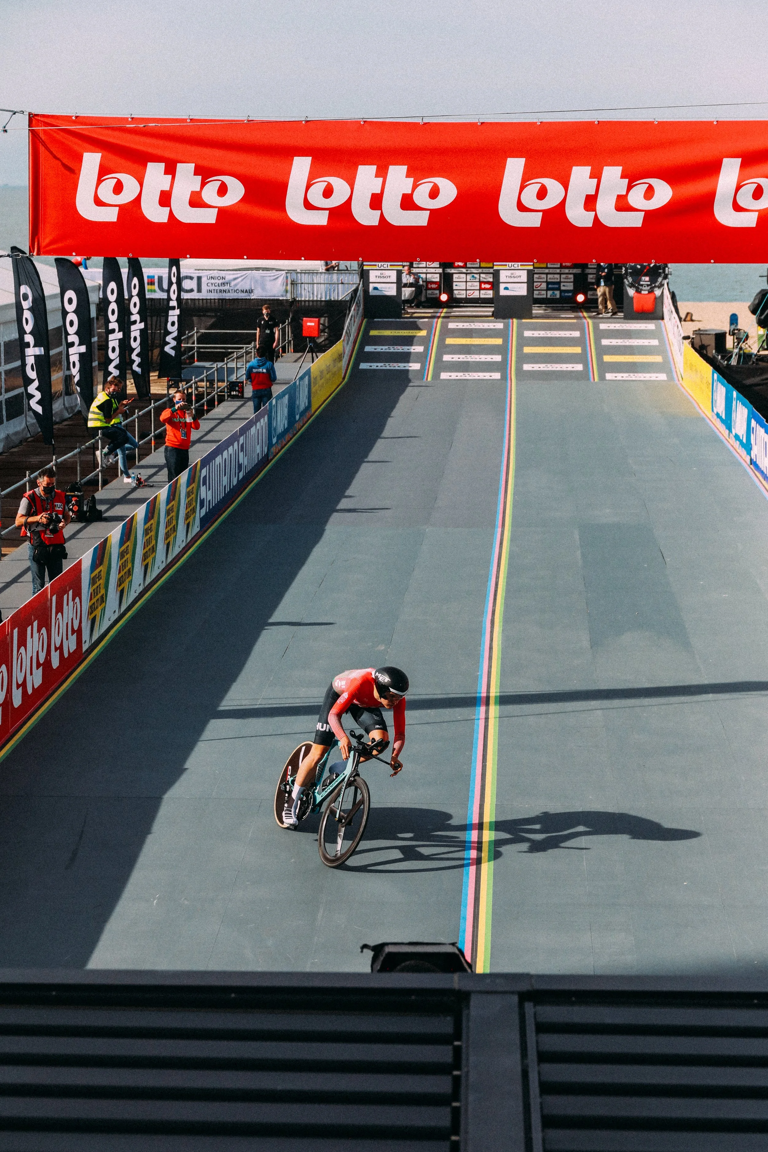 Cyclist in a race on a track under a Lotto banner with crowd and event signage.