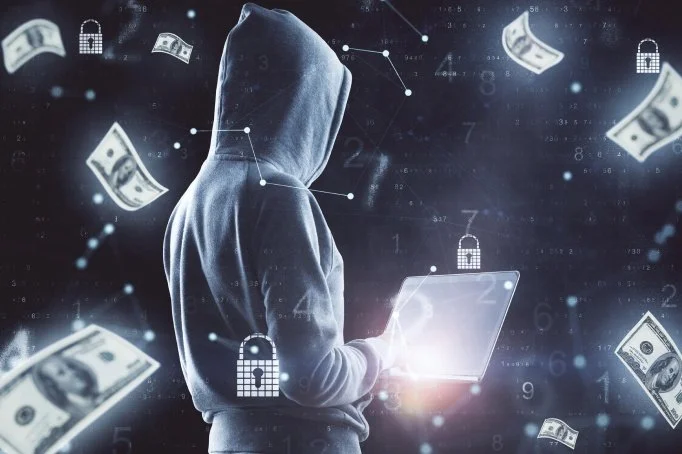 Person in a hoodie holding a laptop, surrounded by flying dollar bills and digital padlocks, symbolizing cybersecurity and online financial theft.