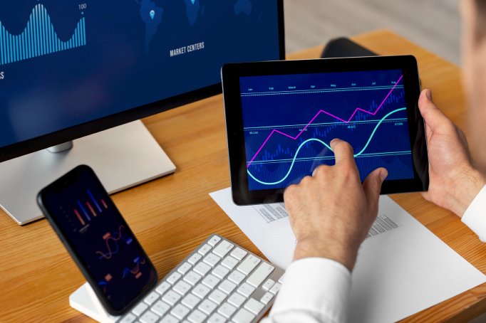 Person analyzing financial charts on a tablet in an office setting, with a computer monitor and smartphone displaying market data.