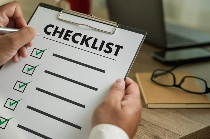Person holding a checklist on a clipboard with checkmarks, sitting at a desk near a laptop and glasses.