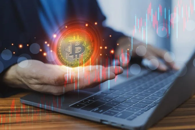 Person using a laptop with a digital Bitcoin logo and financial charts overlaid.