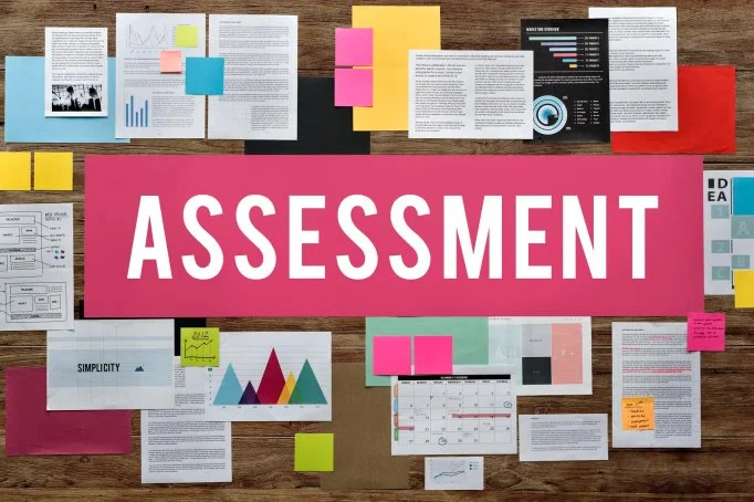 Variety of documents, charts, and sticky notes on a wooden table with a large pink sign that says 'ASSESSMENT'.