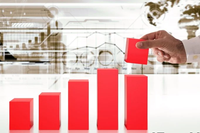 A person in a white shirt places a red block onto a red bar of a rising bar graph, with a digital background of charts and a world map.