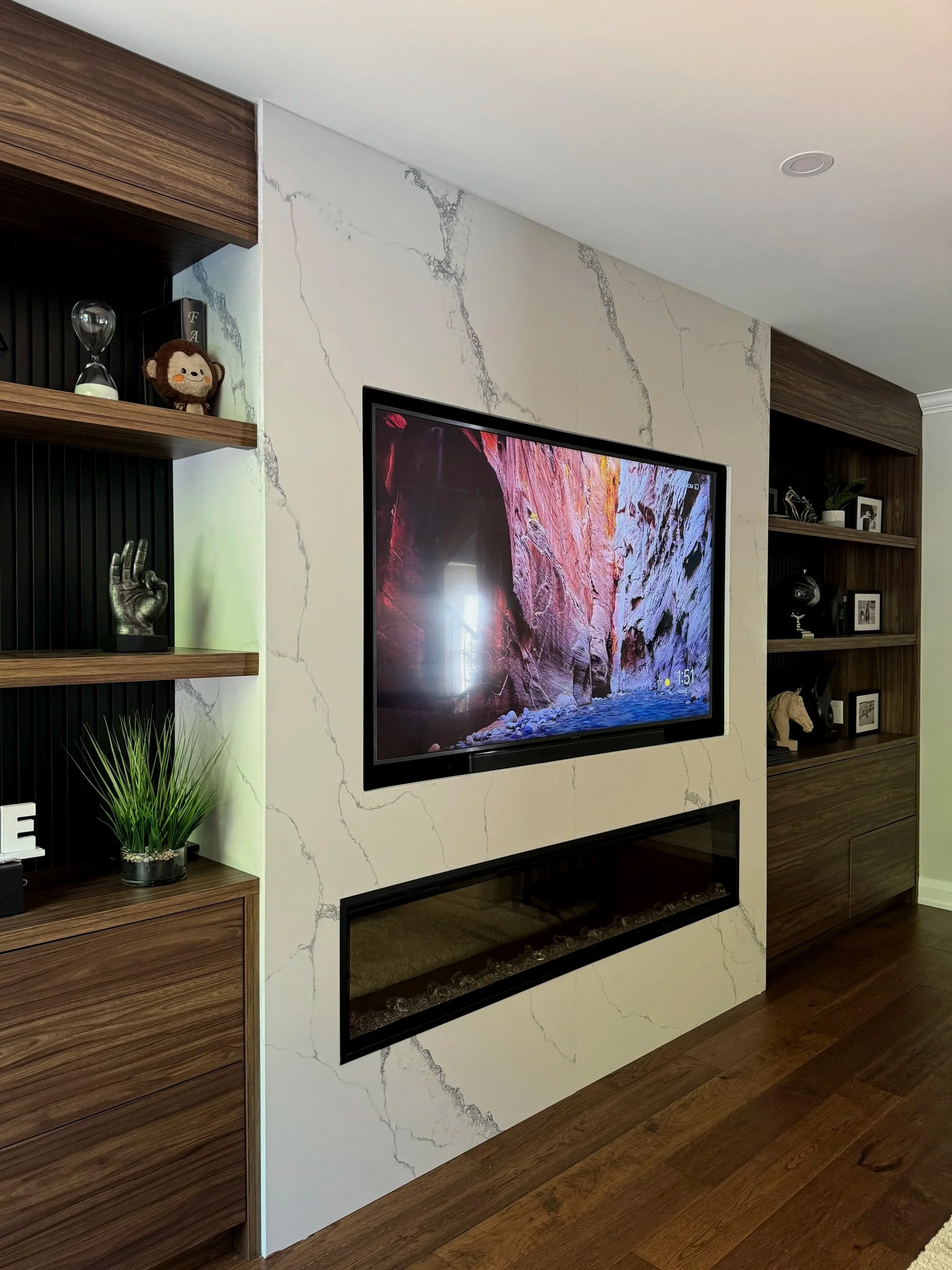 Modern living room with a large wall-mounted TV and electric fireplace. The wall is decorated with white marble-style panels. Wooden shelves display decorative items like a stuffed toy, hourglass, and plants. The floor is dark wood.