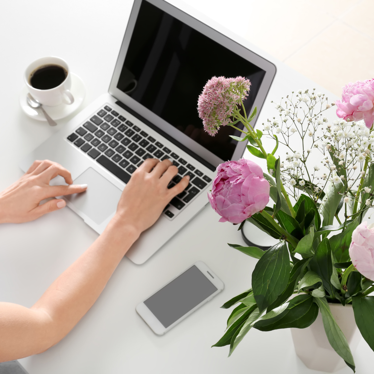 A person typing on a silver laptop with a black keyboard, on a white desk, next to a white smartphone and a cup of black coffee. There is a large bouquet of pink peonies, white baby's breath, and a pink allium in a white vase on the desk.
