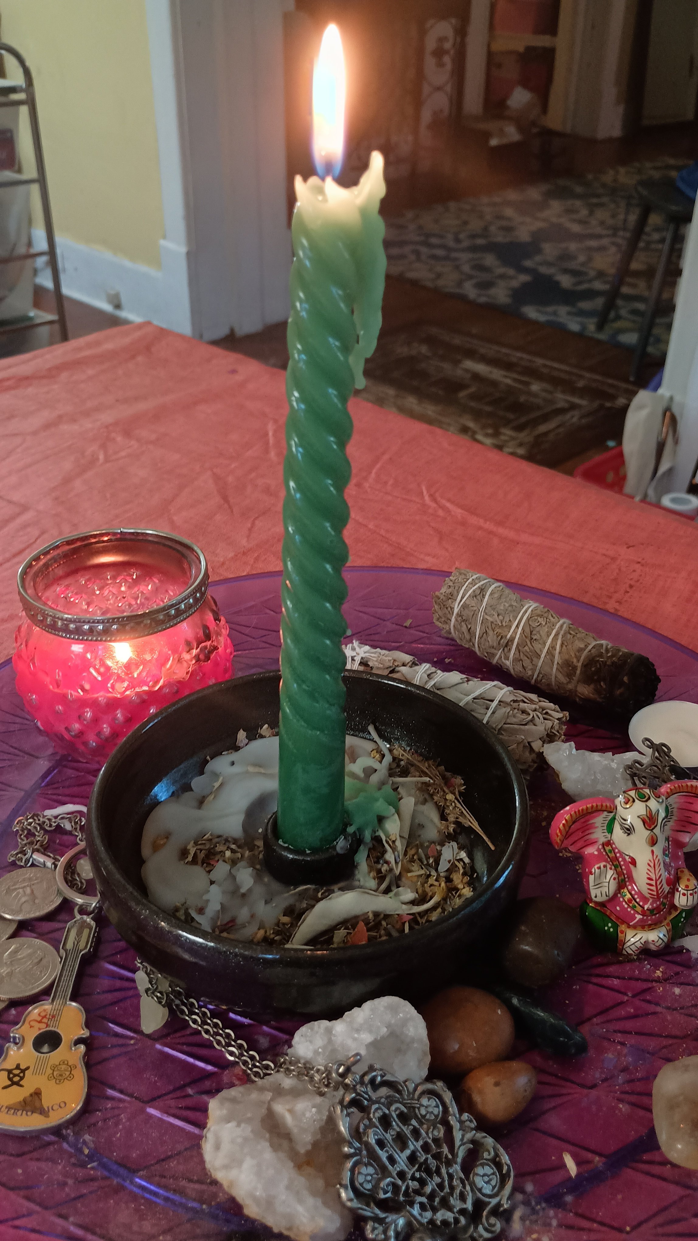 A green twisted taper candle lit with various trinkets, stones, and a small colorful elephant figurine showing a small studio ritual.