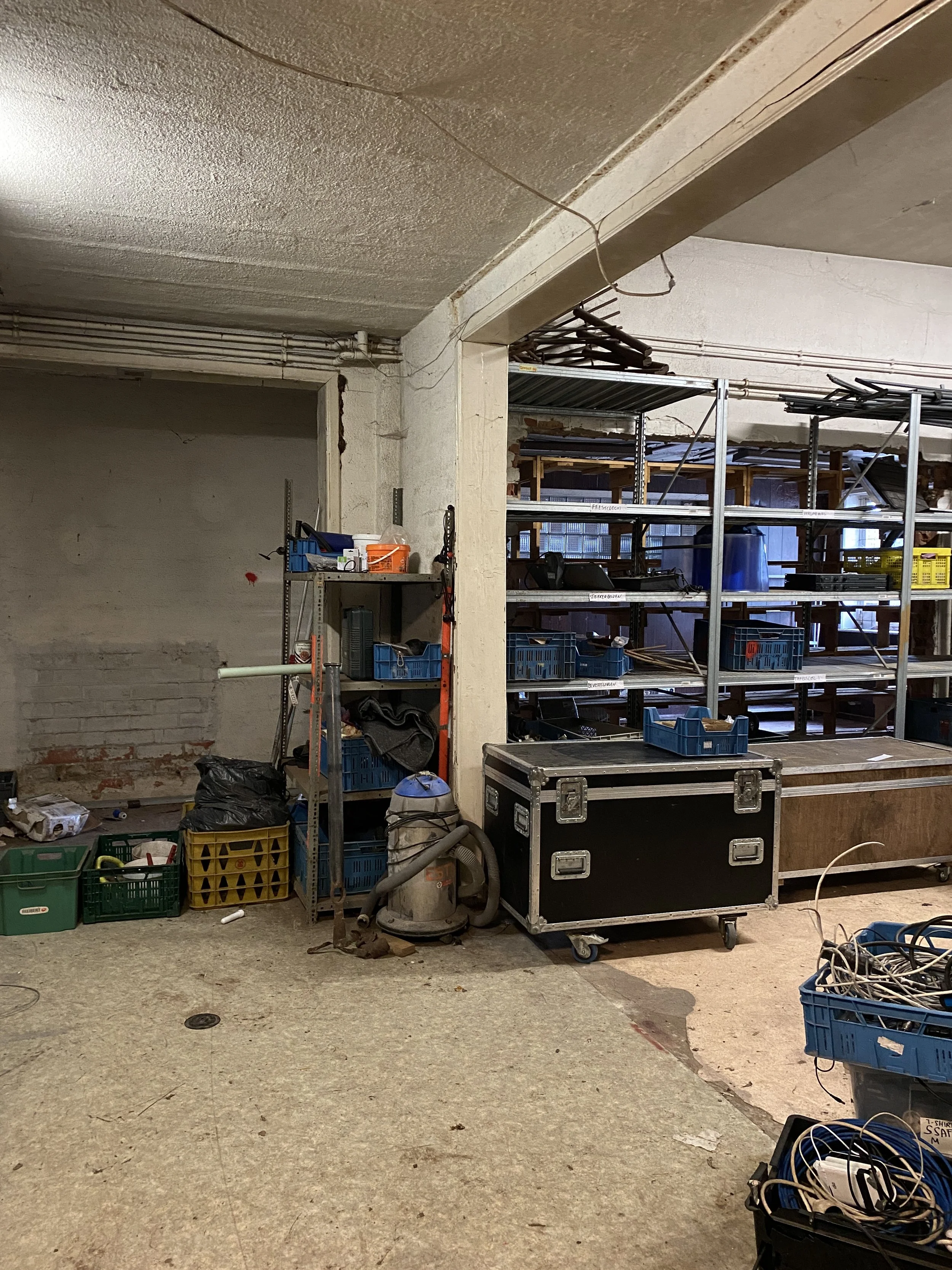 Een binnenruimte met metalen plankenrekken gevuld met diverse bakken en gereedschap. Er staat ook een stofzuiger en andere opslagcontainers.