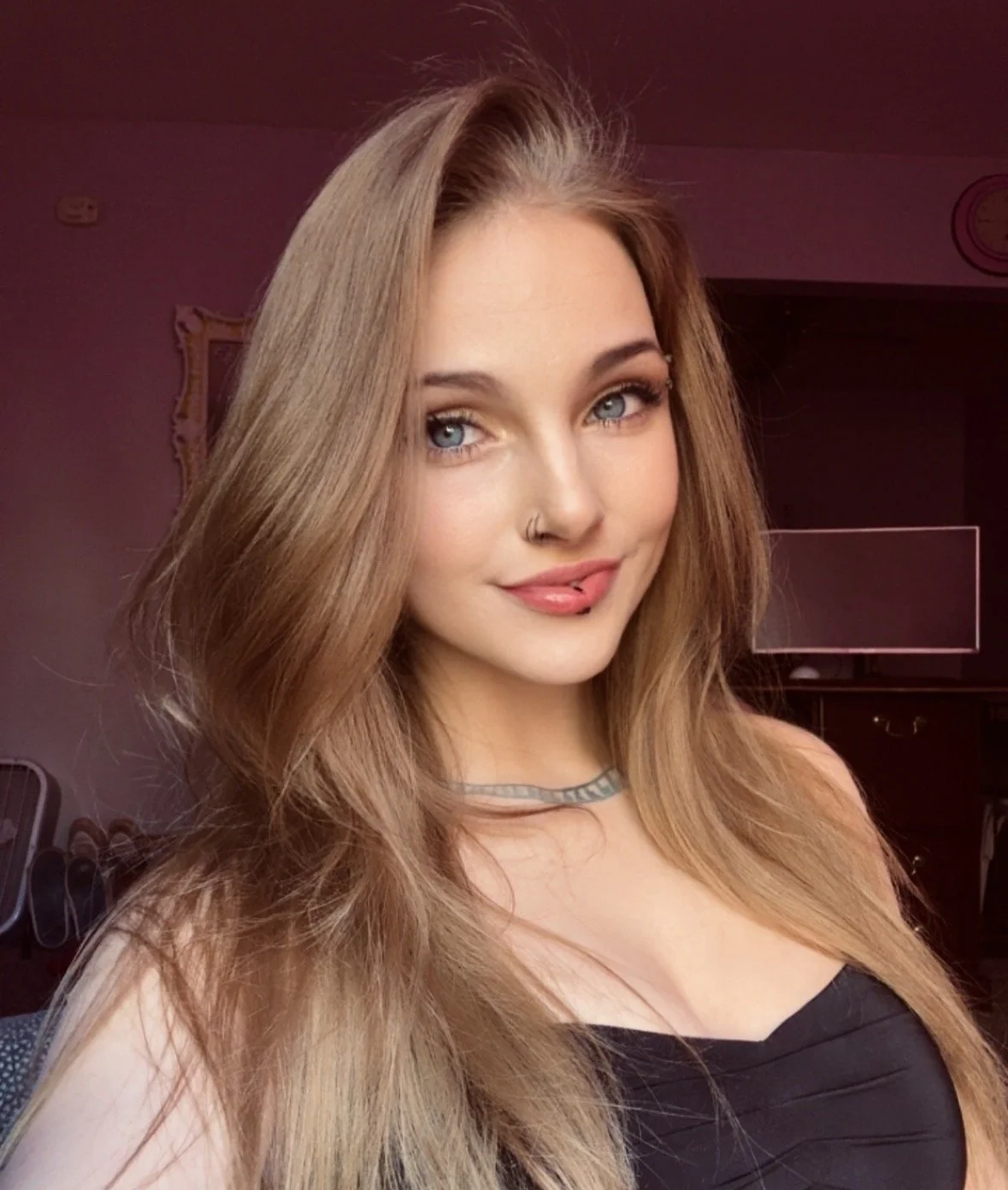 A young woman with long, wavy blonde hair, blue eyes, and a nose piercing, smiling and wearing a black top and a choker necklace, indoors with a purple hue lighting.