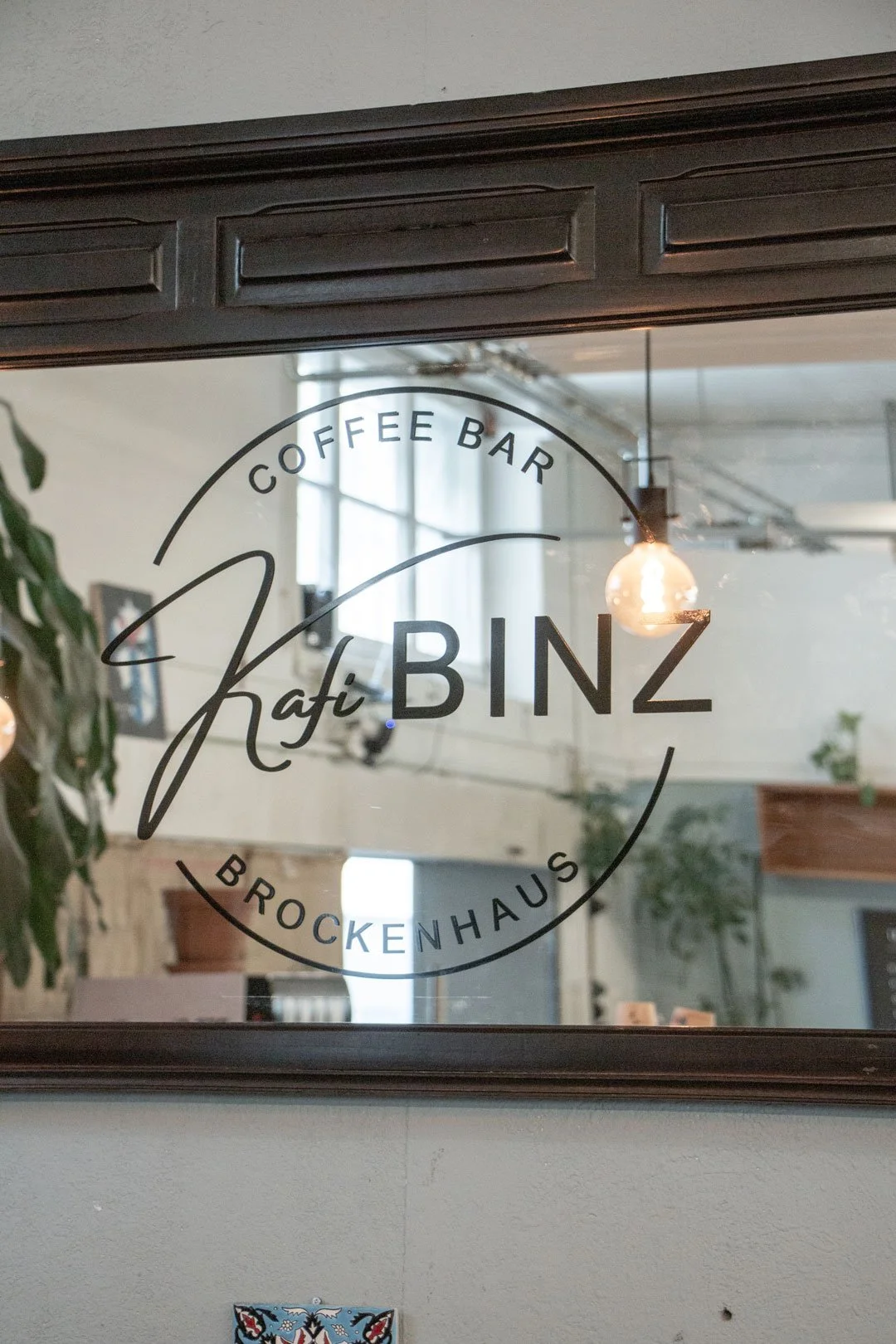 Spiegel mit Siebdruck Aufschrift 'Kafi BINZ Coffee Bar Brockenhaus' in einem Café, Innenraum mit Pflanzen, hellem Licht und Glühbirnen.