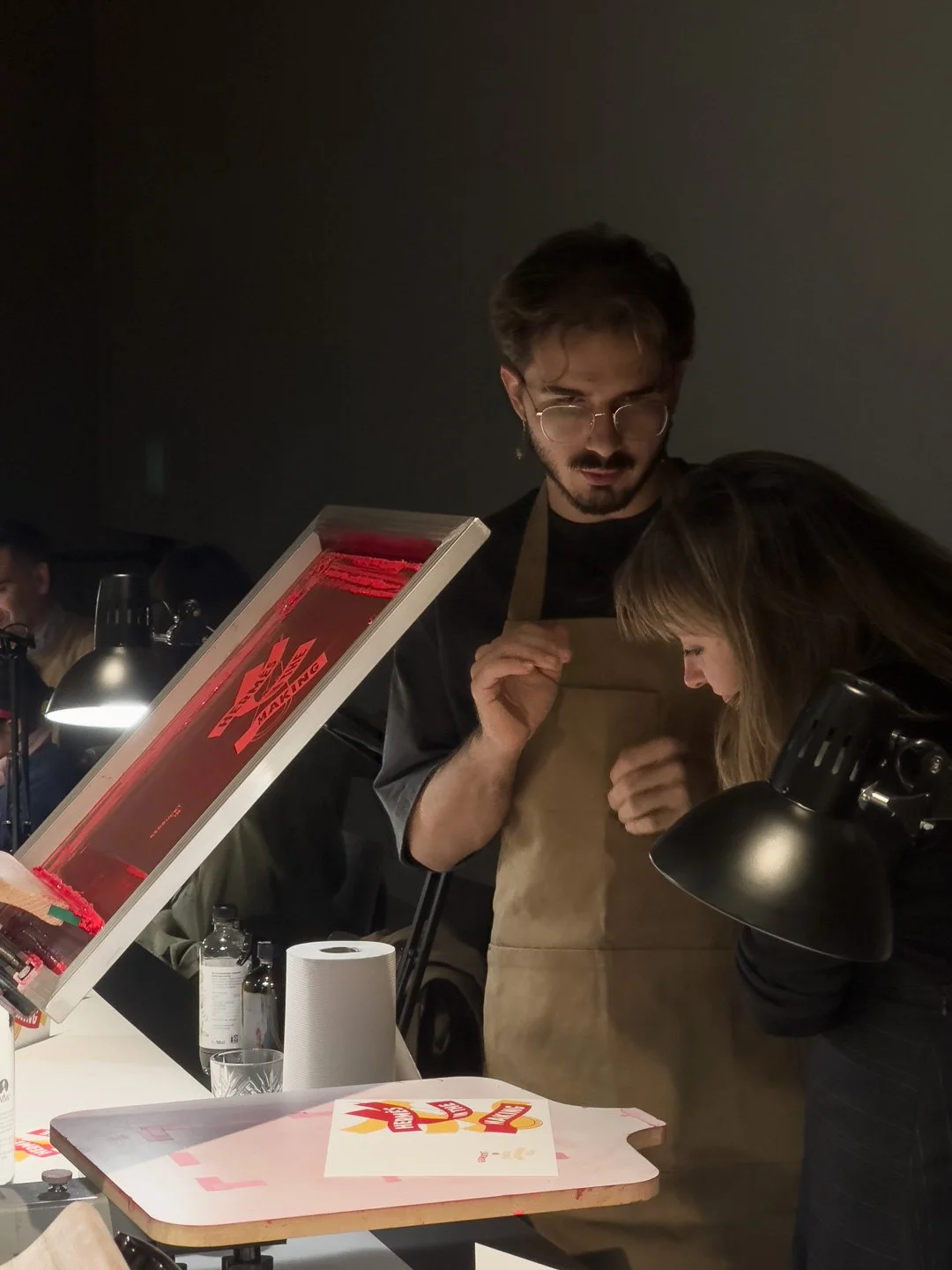 Zwei Menschen, ein Mann mit Brille und ein Mädchen, stehen über einen Tisch mit einem Pizzabrett und einer Pizzaschneidemaschine. Sie tragen Aprons und scheinen bei der Zubereitung oder Präsentation einer Pizza zu sein.