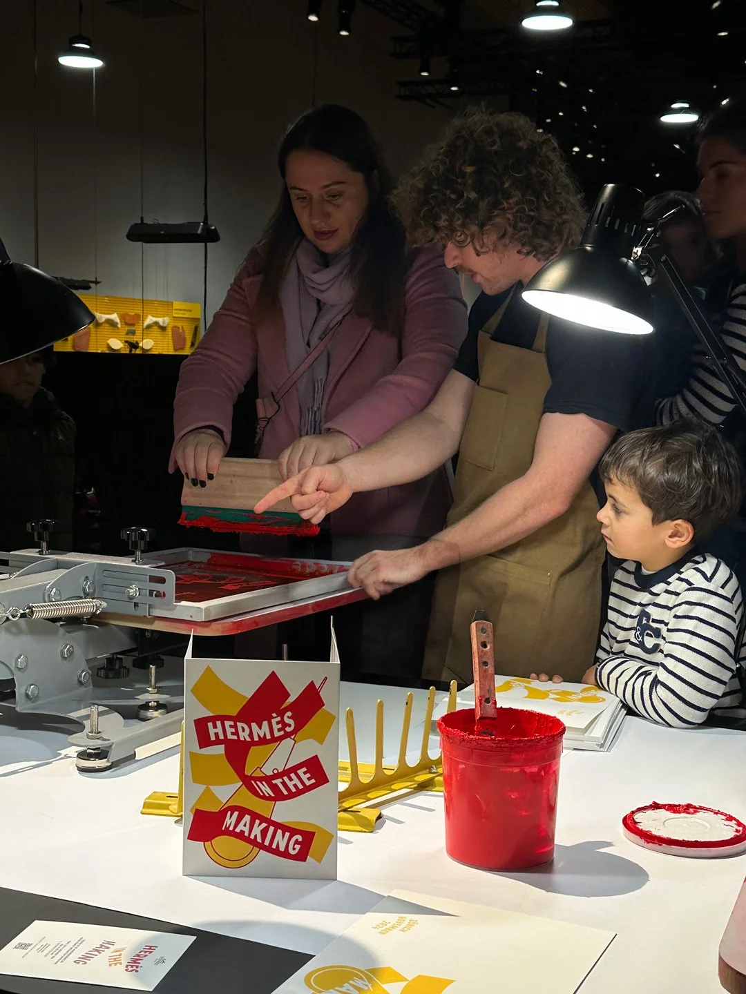 Menschen bei einer Interaktion am Tisch in einer Ausstellung, mit einem Schild, das 'Hermès in the Making' sagt, und einer roten Farbe in einem Behälter.