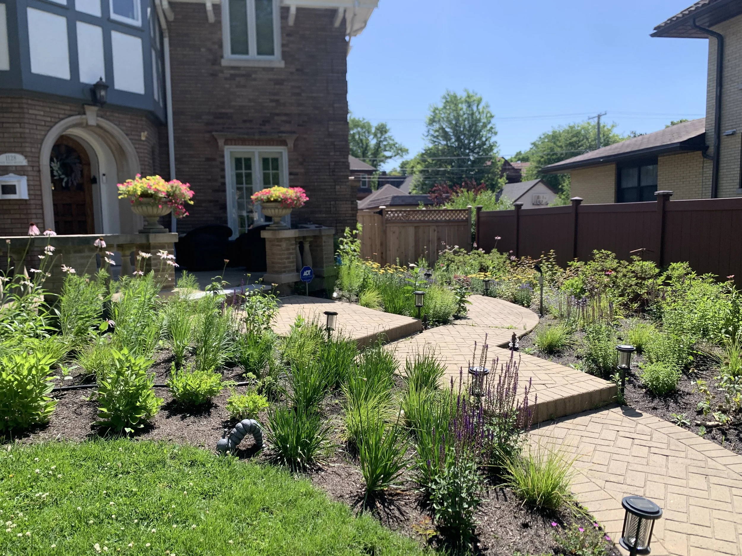 A landscaped front yard Oak Park, Illinois garden with a brick pathway, lush green plants, colorful flowers, and a patio area with potted plants under a blue sky.