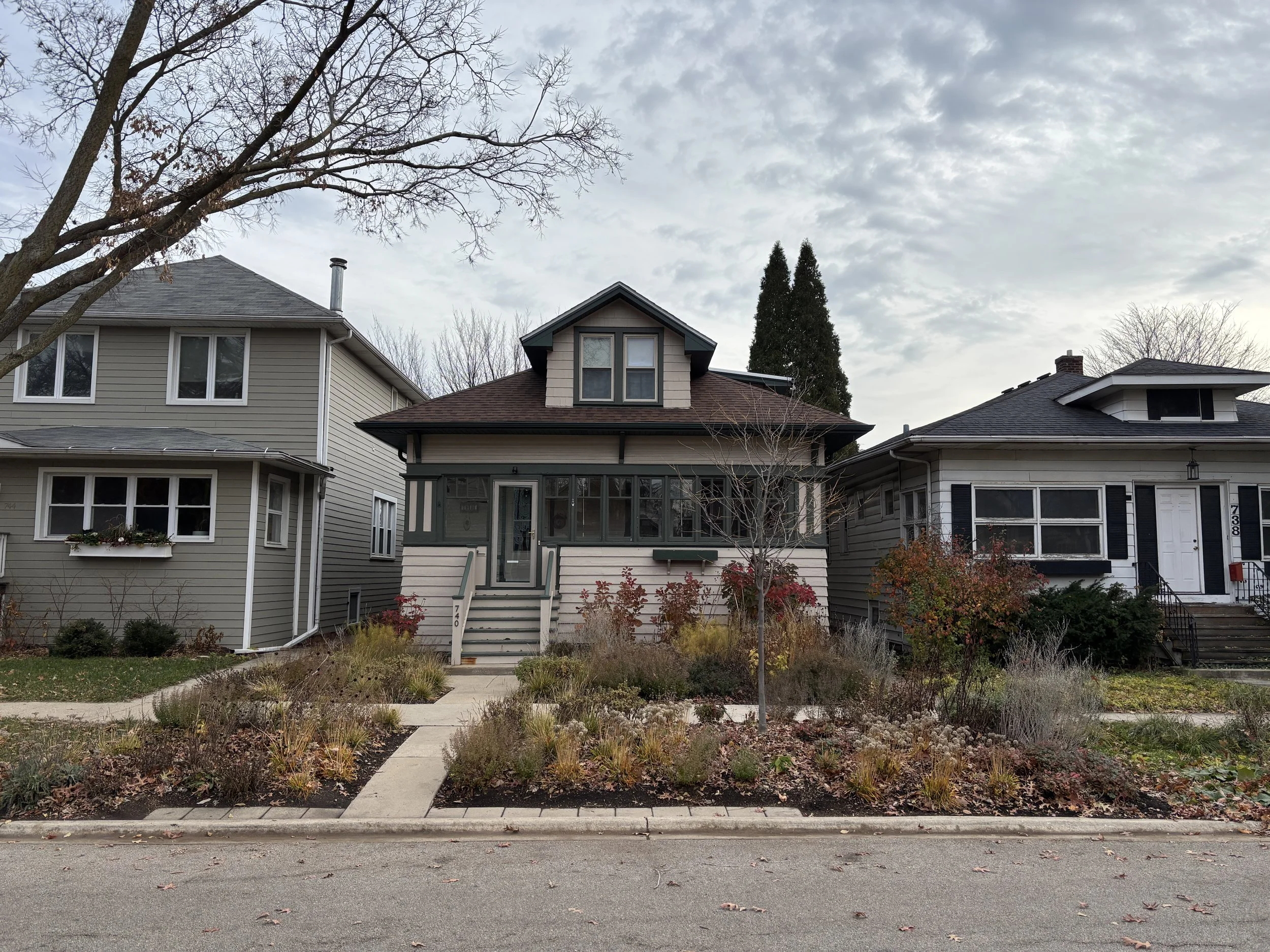 A row of three houses along a street with a garden in front, including a small tree and various plants, under a cloudy sky.