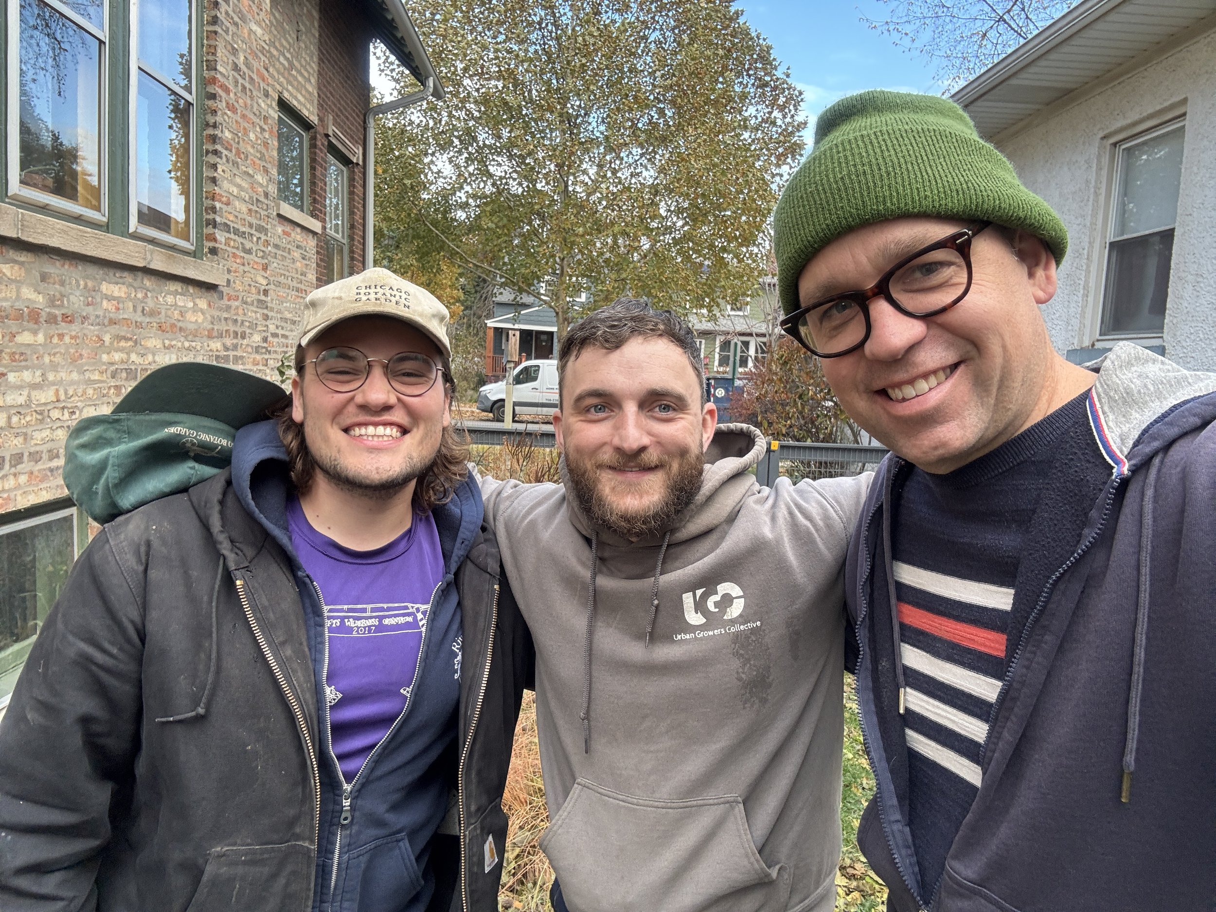 Three men smiling and taking a selfie outdoors in a neighborhood with houses and trees in the background during fall.