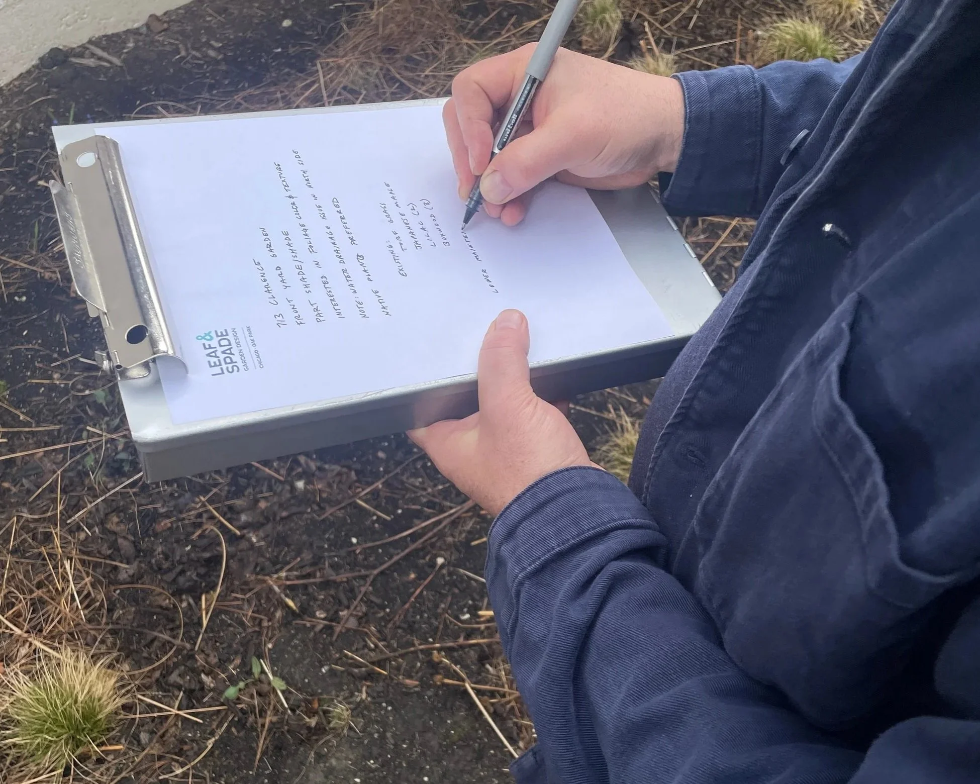 Person wearing a dark denim jacket writing on a clipboard with a silver pen outdoors on a patch of dirt and sparse grass.