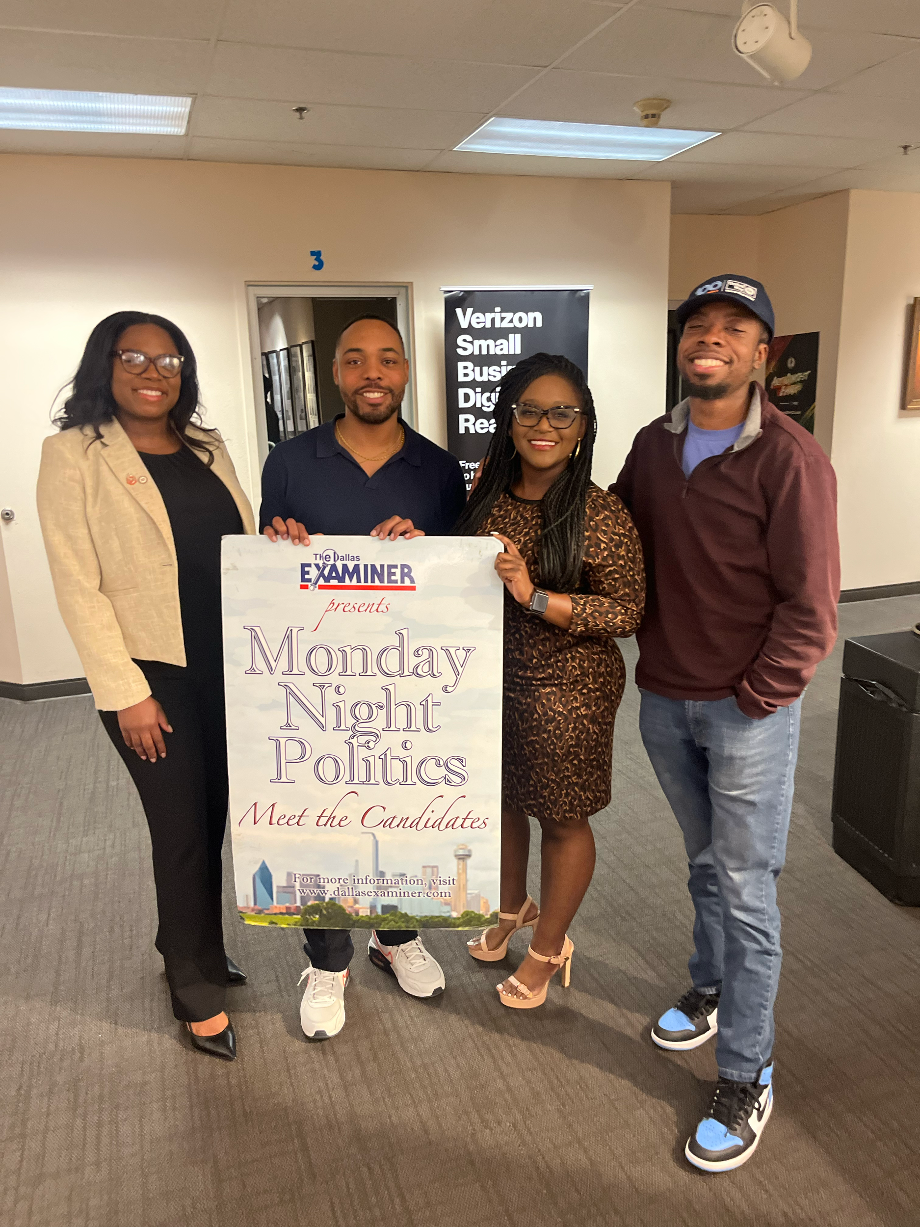 Group of four people posing with a "Monday Night Politics" event poster in an office setting.