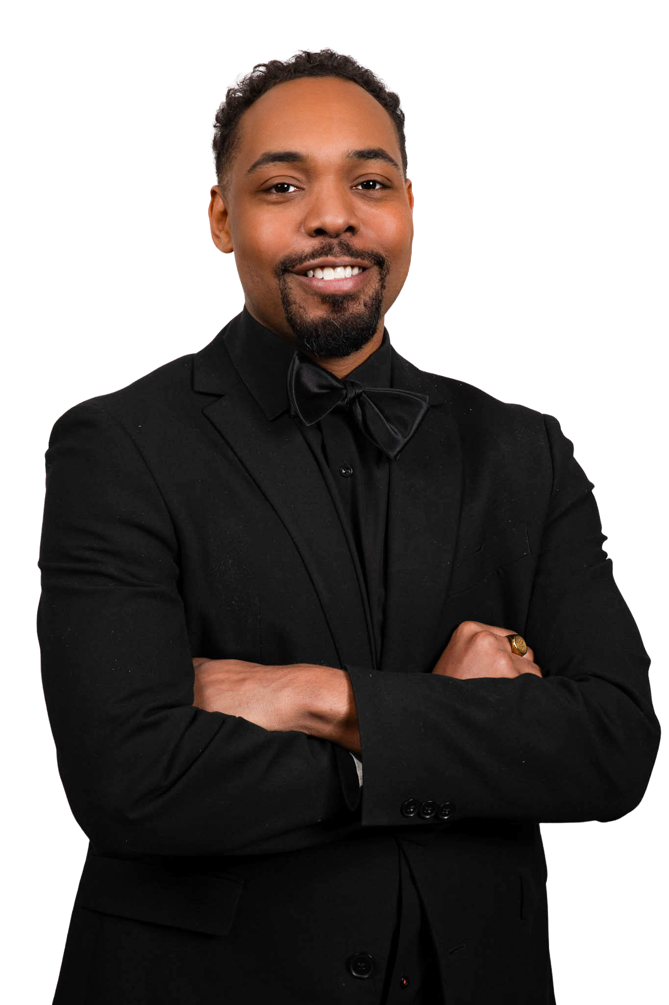 Smiling man in formal black tuxedo with a bow tie, crossed arms, wearing a gold watch, set against a plain beige background.