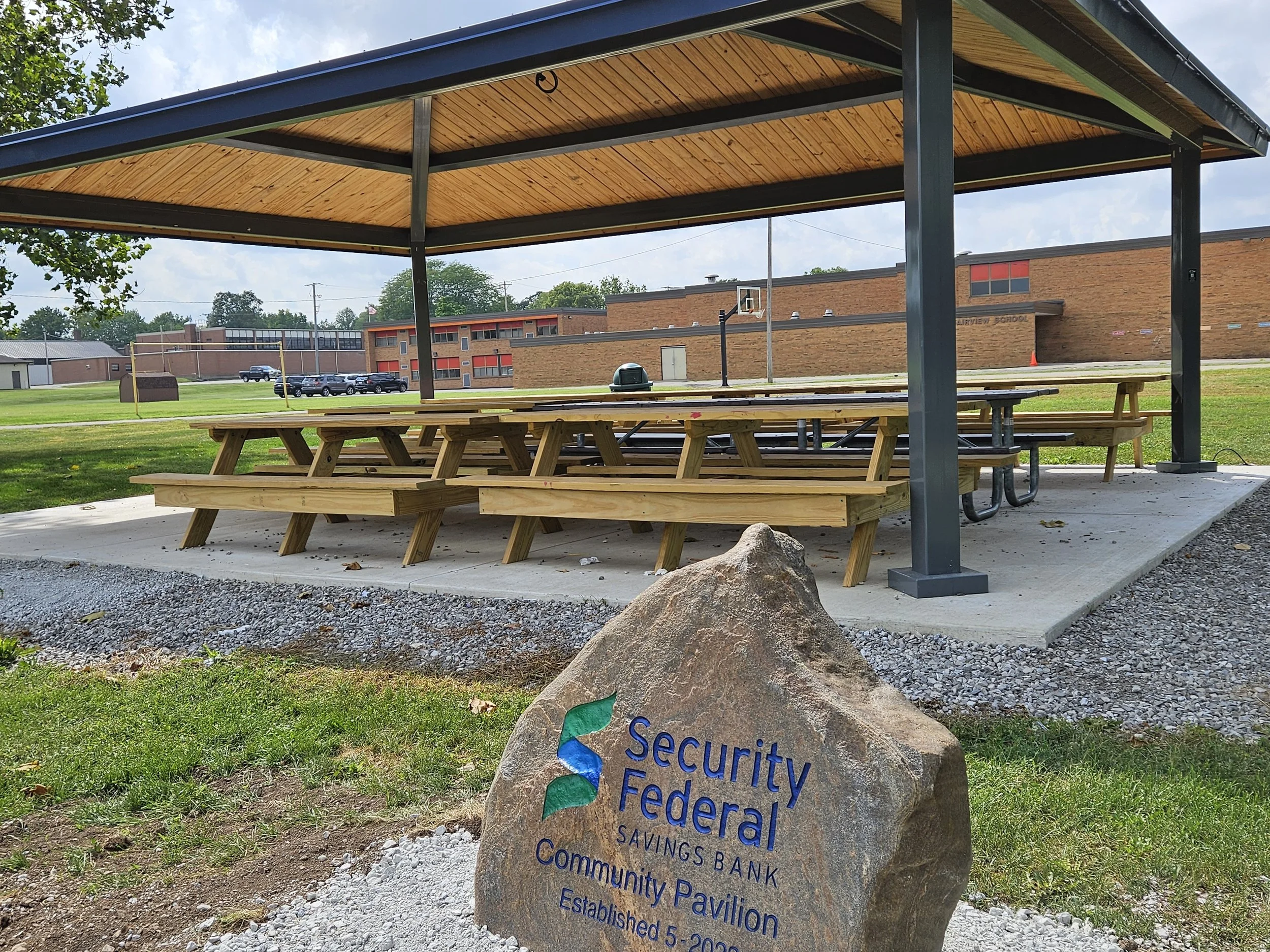 Image contains a metal and wooden awning with picnic tables. Fairview School is in the background, and in the foreground is a stone sign that reads: "Security Federal Savings Bank Community Pavilion".