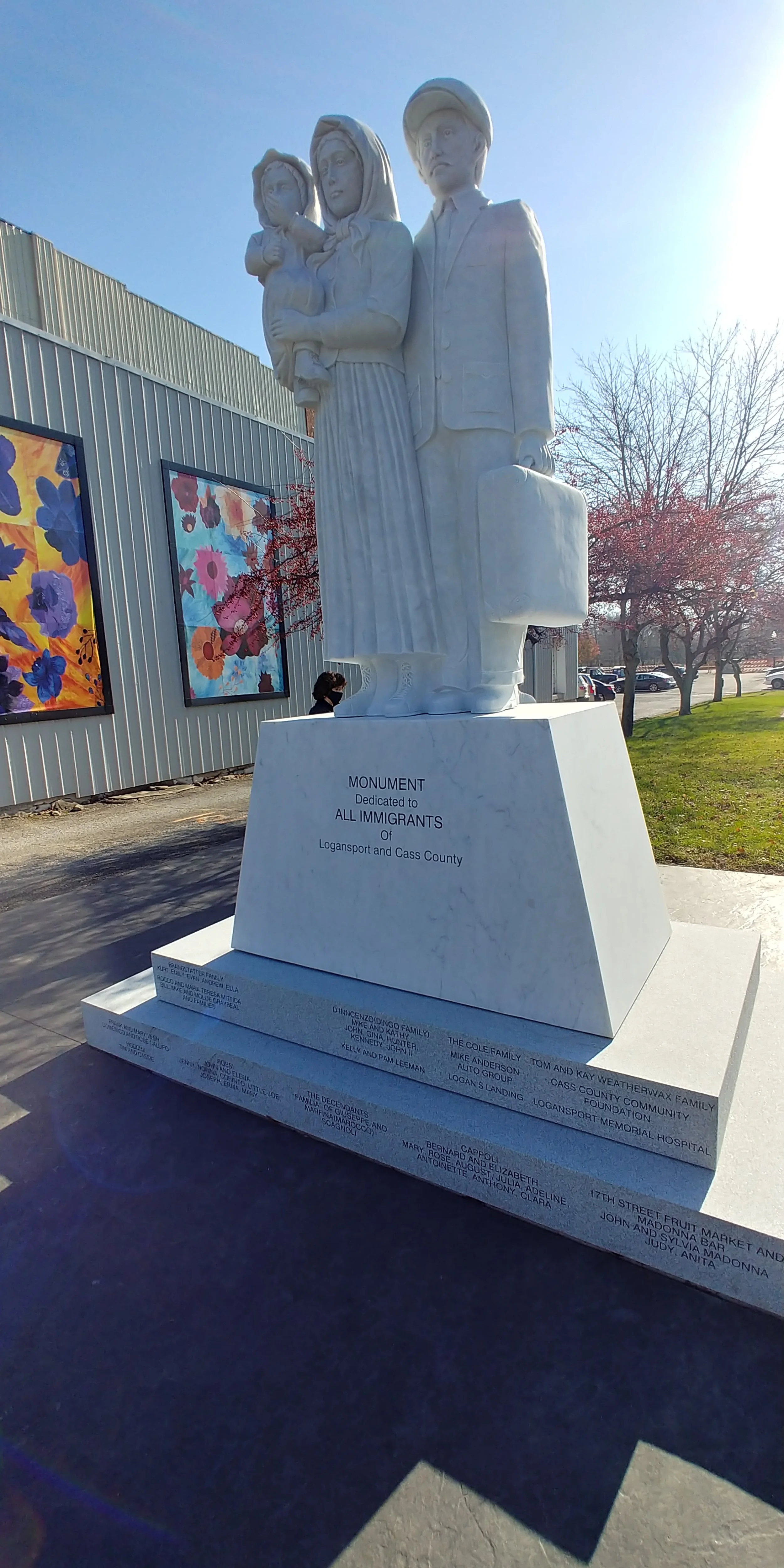 Image contains a white statue of a couple carrying bags and a baby. Sign reads: "Monument dedicated to all immigrants of Logansport and Cass County."