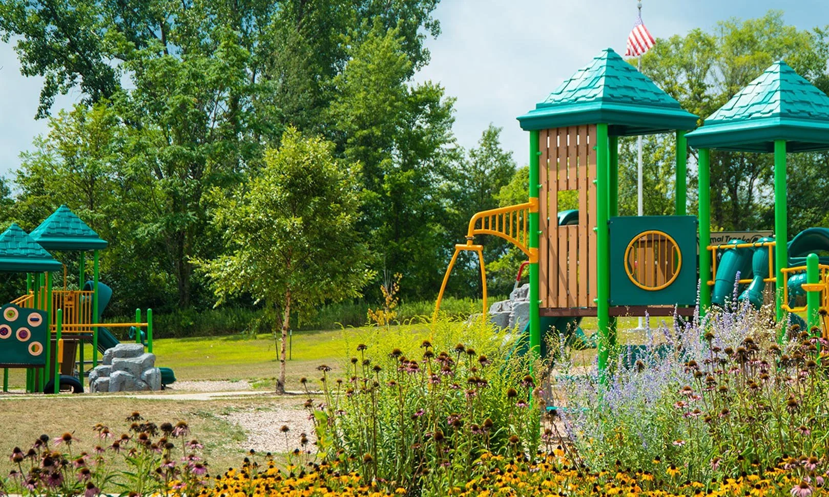 Image contains a green and teal playground structure with flowers in the foreground.