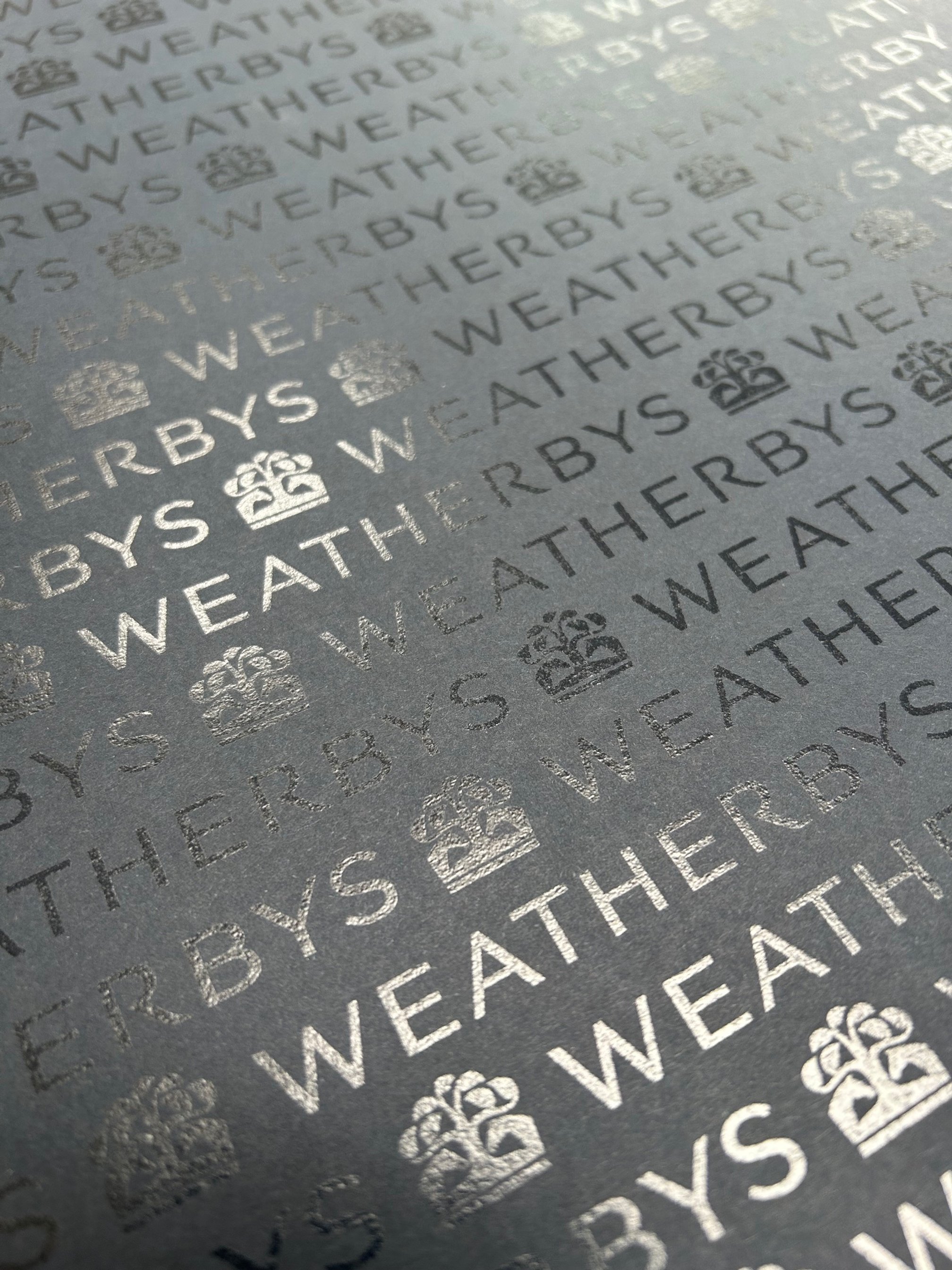 Close-up of black textured paper with a repeating pattern of the word 'WEATHERBYS', a crown icon, and the letter 'W' in silver print.