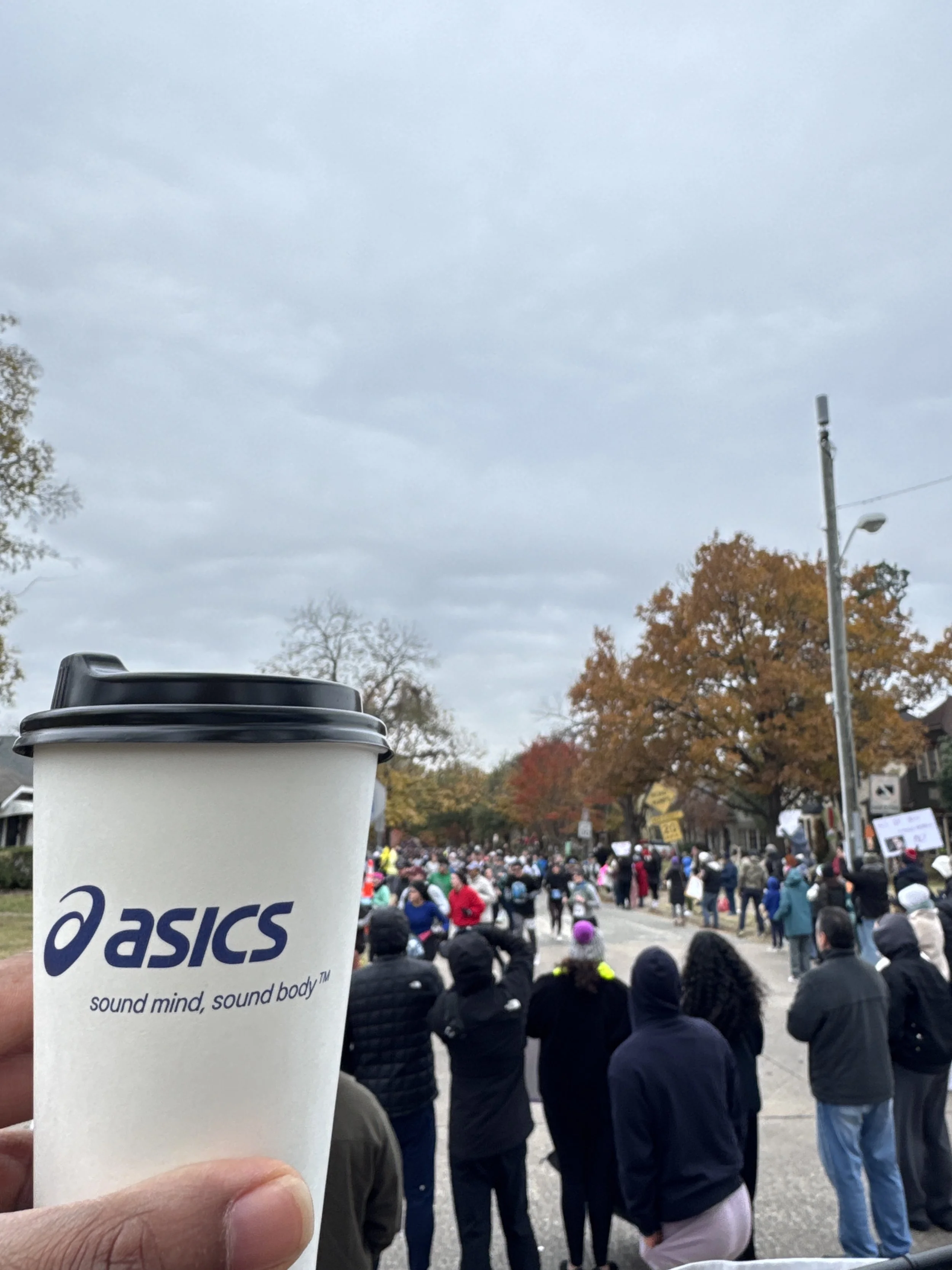 A person holding a white Asics coffee cup in front of a crowded street protest with many people, some holding signs, in fall with orange and yellow trees and cloudy sky.