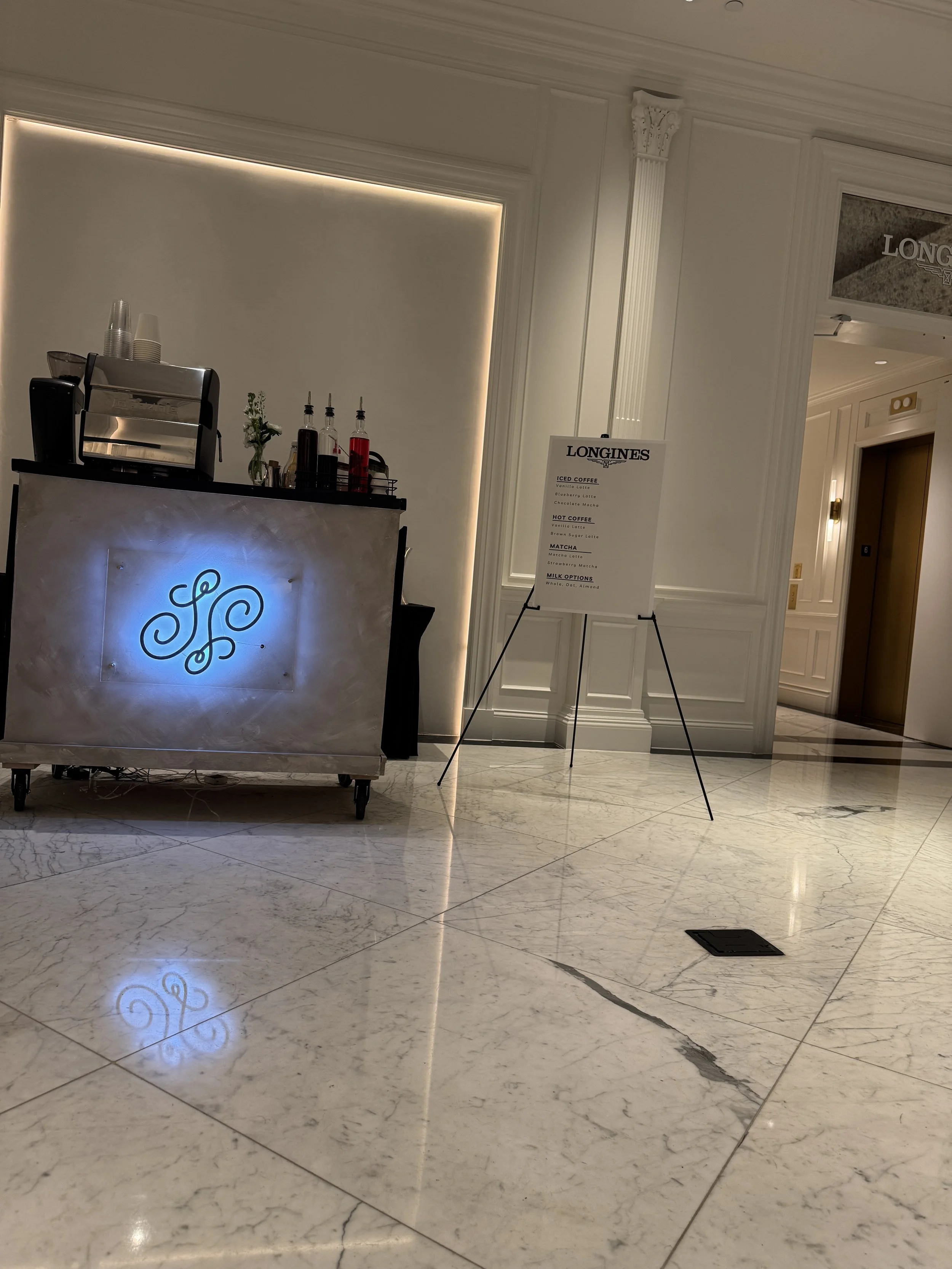 A small coffee stand in a luxury hotel lobby with a neon sign on the front, bottles of syrup on the counter, and a signboard listing coffee options.