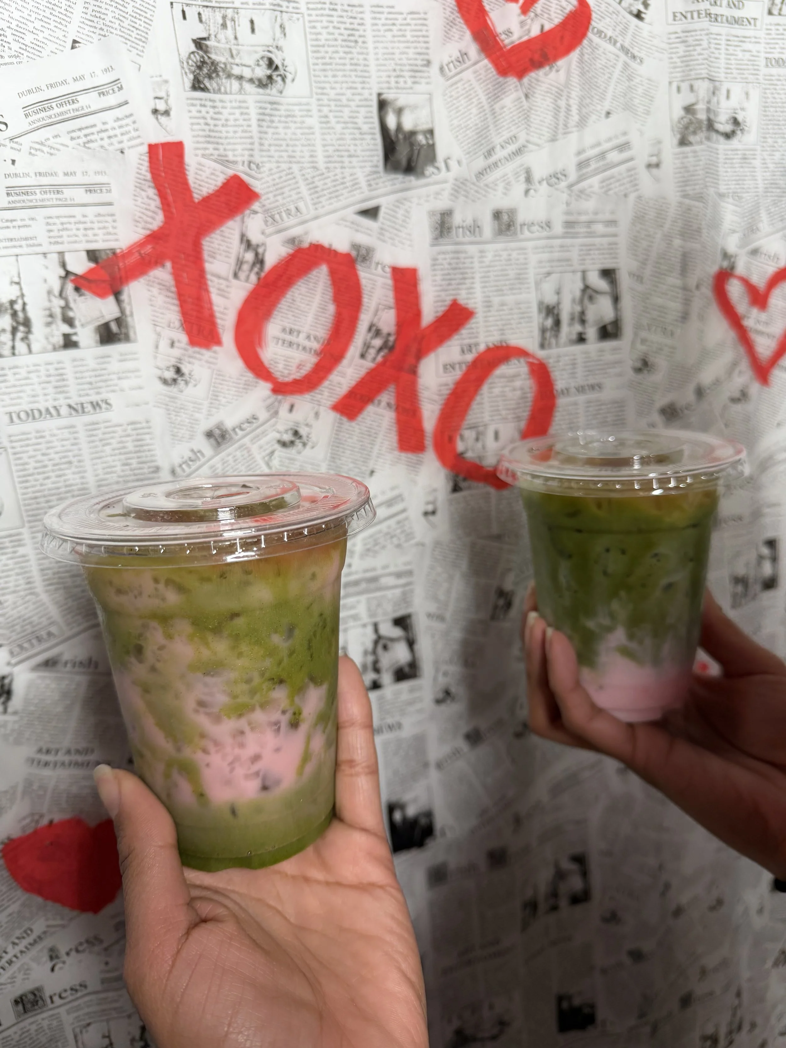 Two hands holding cups with green and pink swirl drinks against a newspaper-themed wall with red graffiti.