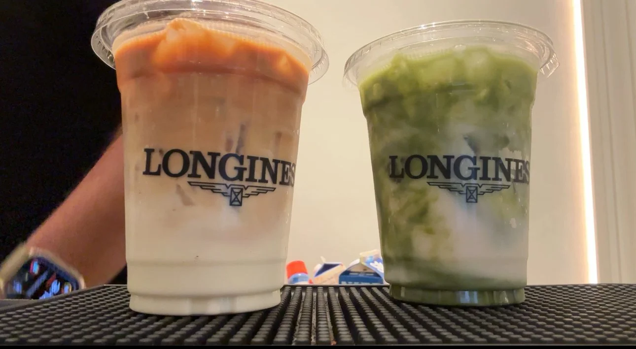 Two plastic cups with the LONGINES logo on them, one containing a layered drink with orange and cream colors, and the other containing a green and white drink, placed on a textured black surface with a person's arm and a watch in the background.